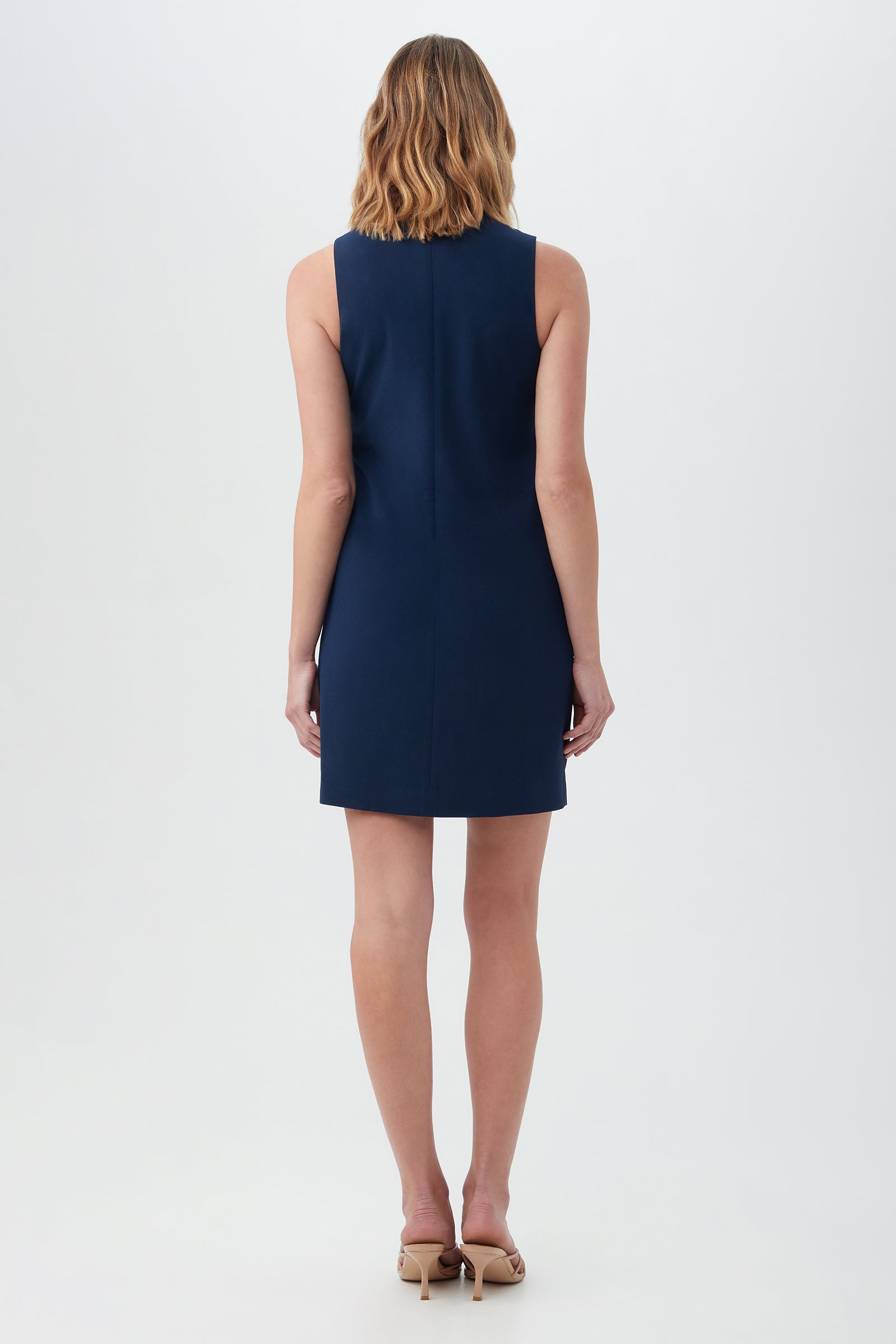 A woman with shoulder-length, wavy hair stands facing away, wearing the Town N Country Dress—a sleeveless, knee-length navy blue dress—and nude heels against a plain light background.