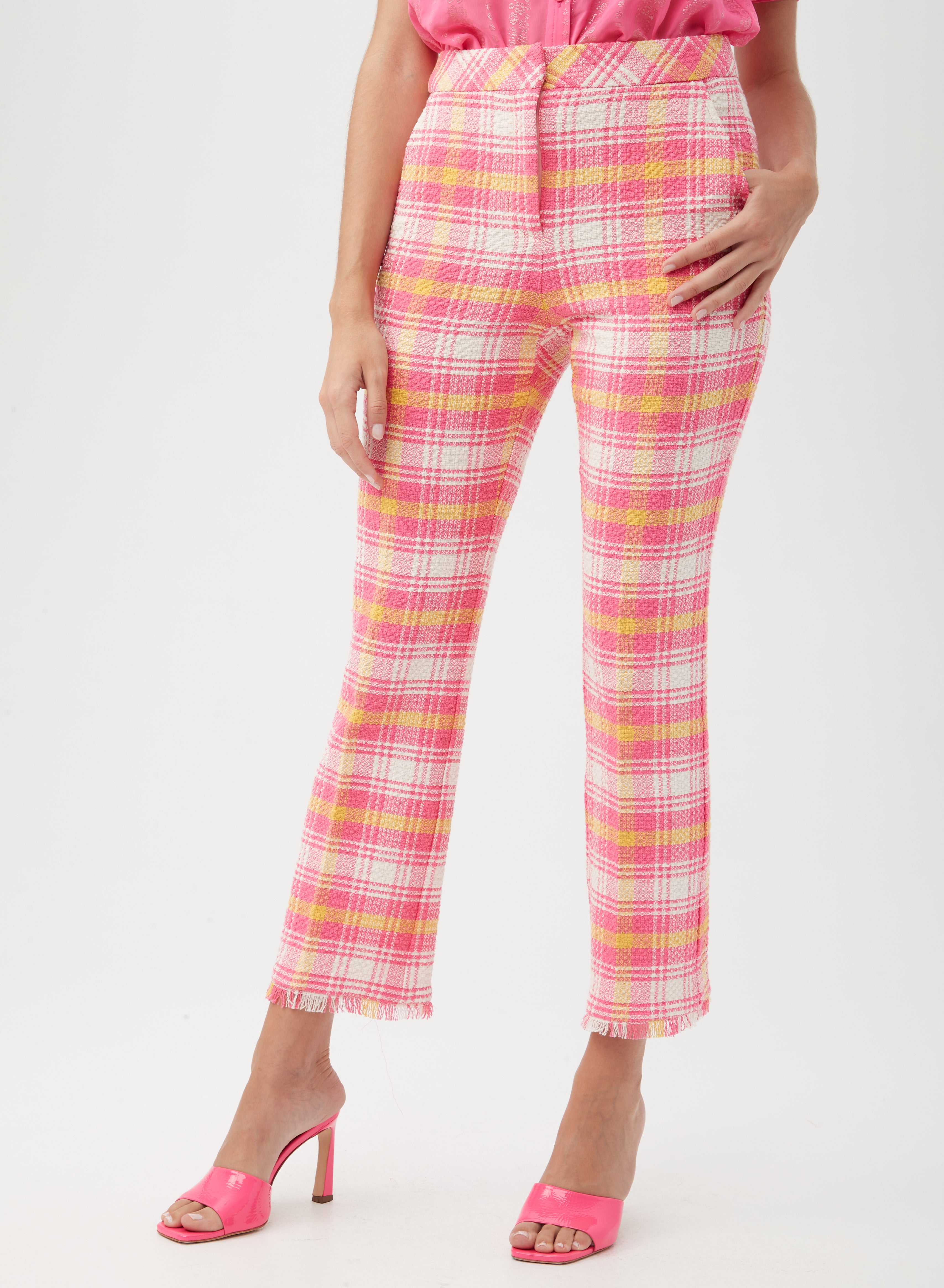 Someone wears the Lulu Fringe Pant in a multi-color plaid design with frayed hems, paired with a pink top and bright pink open-toe heeled sandals, standing against a plain background.