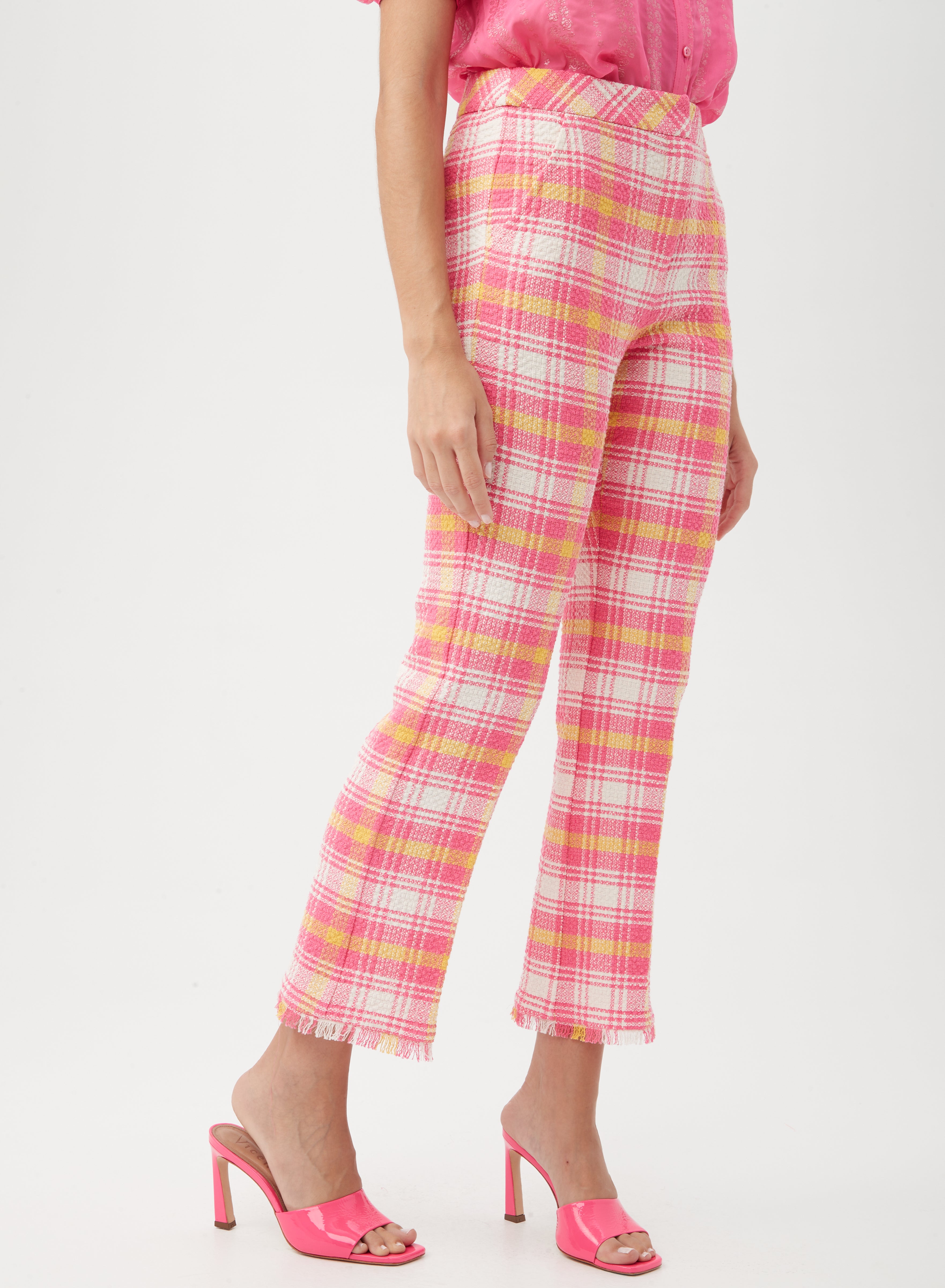 A person wears the Lulu Fringe Pant, high-waisted with pink and yellow plaid and frayed hems, paired with a pink shirt and bright pink heeled sandals. The image captures the outfit from the waist down against a plain background.