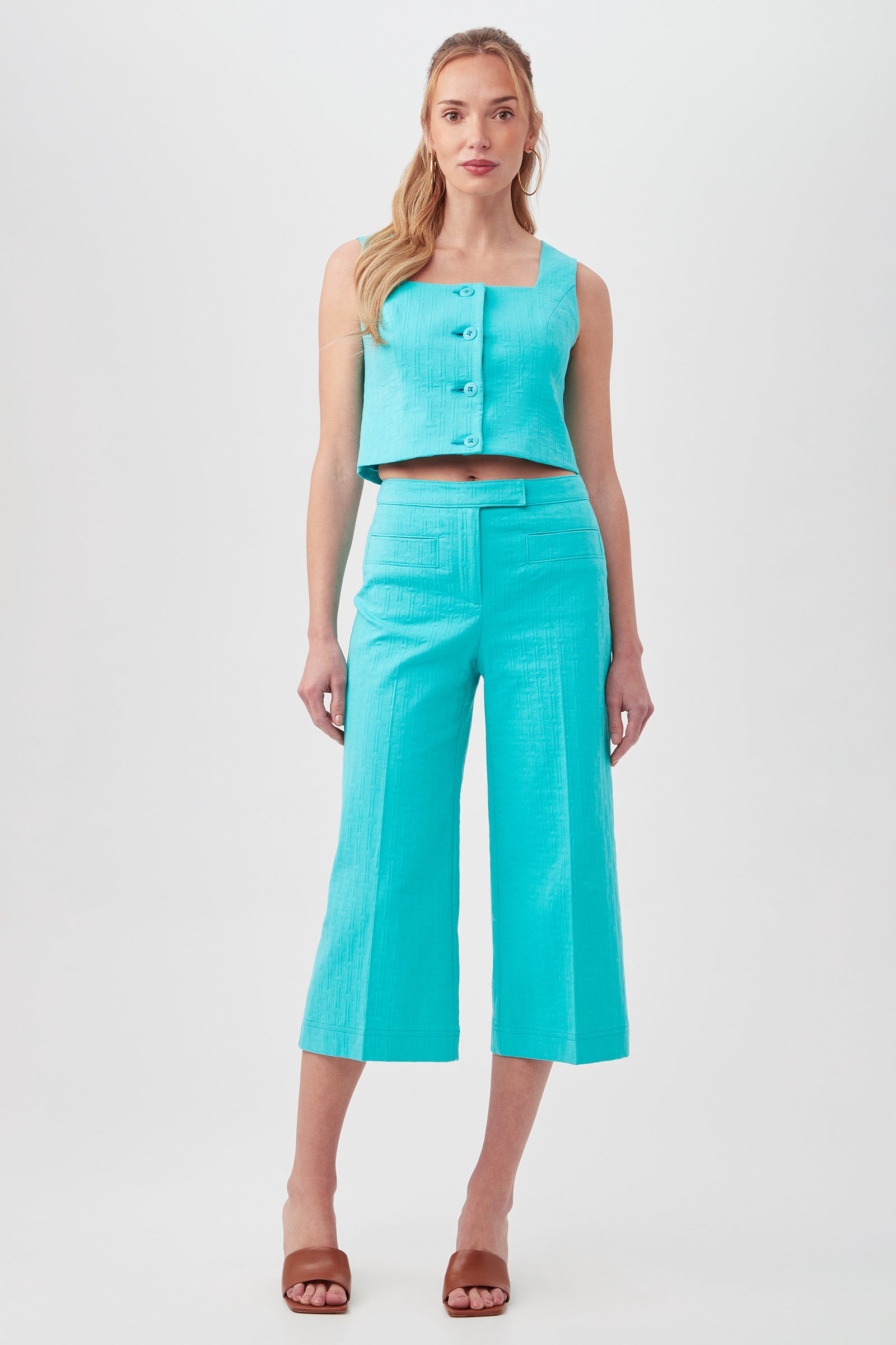 A woman with long blonde hair stands facing forward against a white background, wearing the Refreshment 2 Top— a sleeveless turquoise crop top with large buttons—paired with matching high-waisted, wide-leg cropped pants and brown heeled sandals.