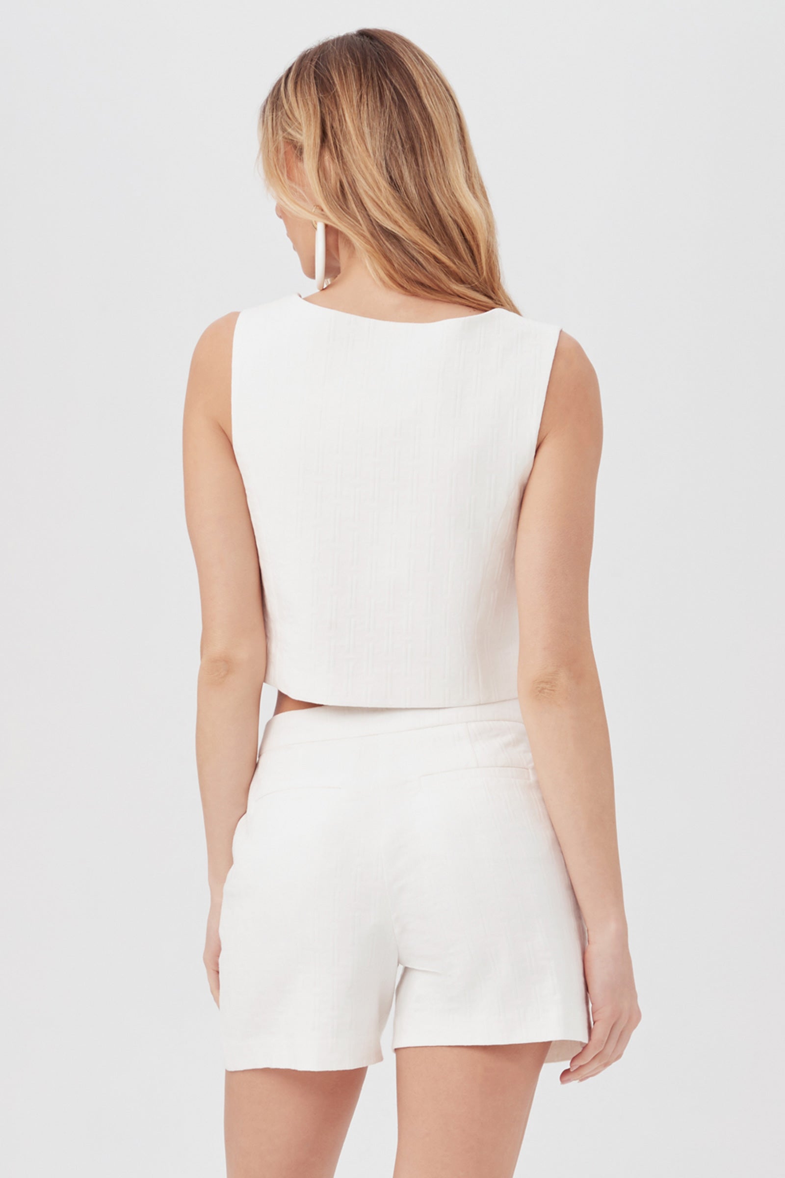 A woman with long, light brown hair stands with her back to the camera, modeling the sleeveless white Refreshment 2 Top paired with matching high-waisted shorts against a plain light background.