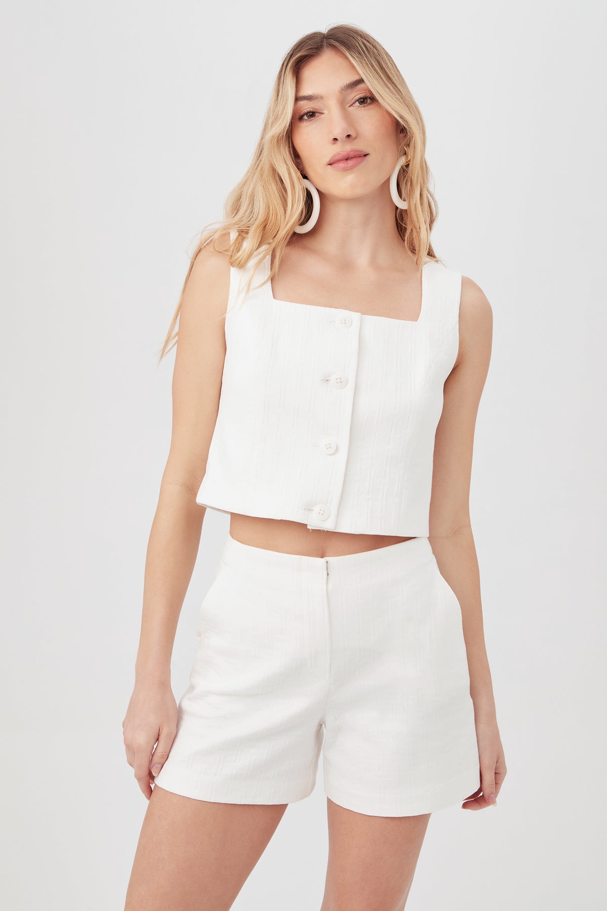 A woman with long, wavy blonde hair wears the Refreshment 2 Top in Whitewash, a sleeveless white button-up crop top, posing against a plain light background.