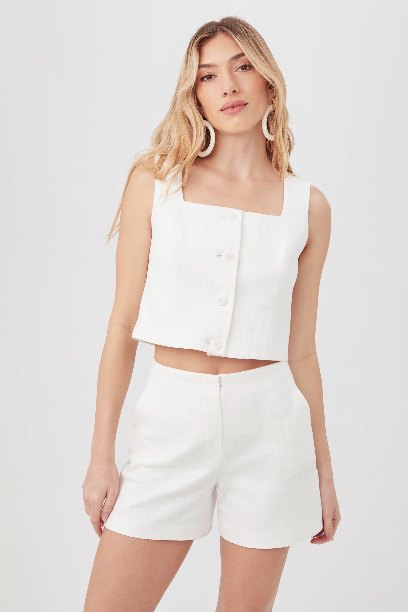 A woman with long, wavy blonde hair wears the Refreshment 2 Top in Whitewash, a sleeveless white button-up crop top, posing against a plain light background.