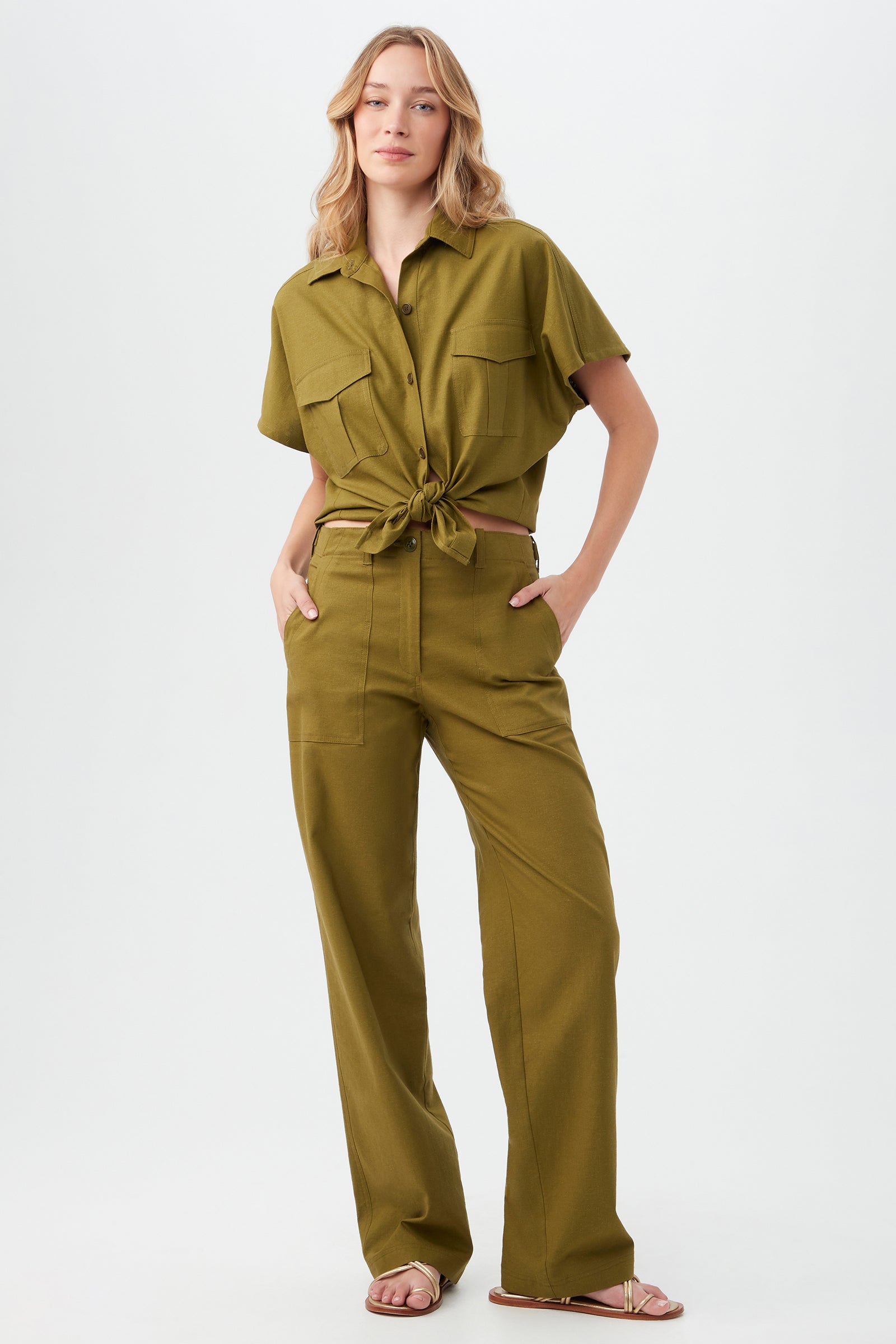 A woman with long wavy blonde hair stands with hands in her pockets, wearing the Antonia Pant—olive green, high-waisted, wide-leg—and a matching button-up shirt tied at the waist. She completes the look with sandals against a plain white background.