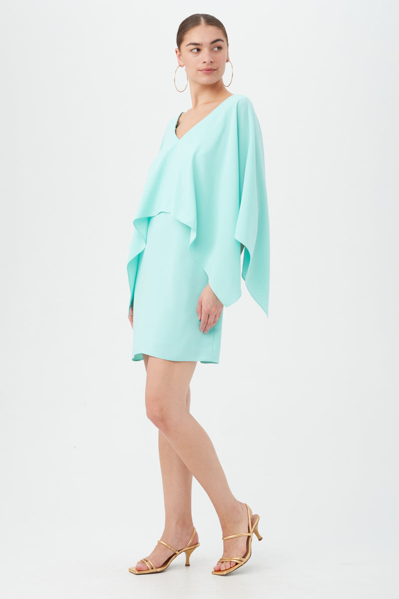 A woman poses against a white background wearing the Azzurra Dress, a light blue, loose-fitting style with wide sleeves, paired with gold strappy heels. Her hair is tied back and she accessorizes with large hoop earrings.