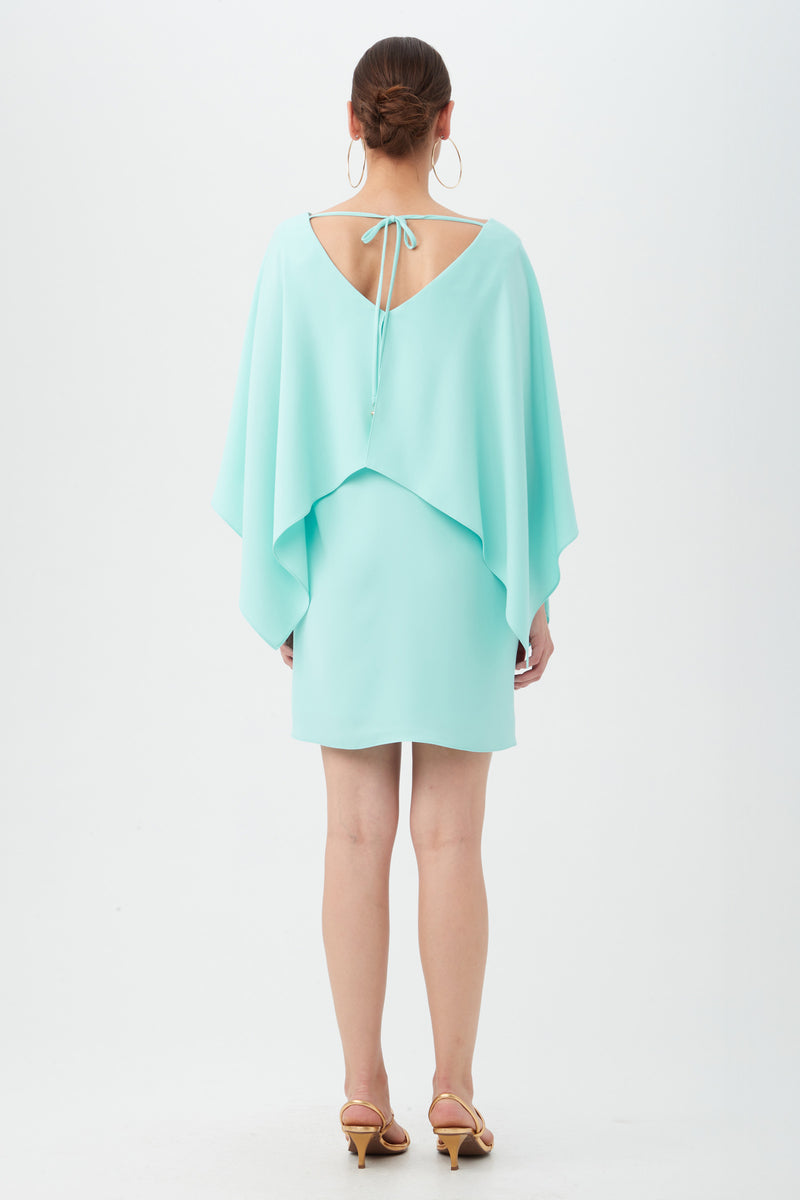 A woman with her hair in a bun, large hoop earrings, and beige heels is seen from the back wearing the Azzurra Dress—a light blue style featuring a cape-like overlay and V-shaped open back tied with a string.
