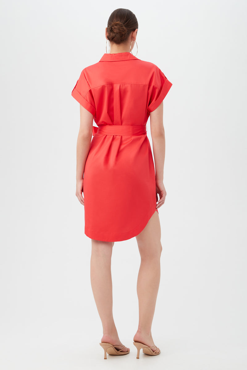 A woman stands facing away, wearing the short-sleeved, belted red Simpatico Dress and beige sandals. Her hair is tied back in a neat bun as she poses against a plain white background.