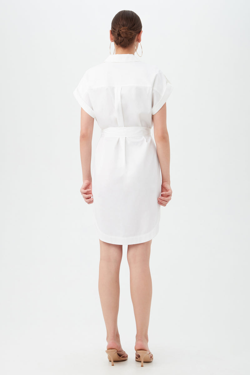 A woman with her hair in a low bun faces away, wearing the Simpatico Dress—a white, short-sleeve, knee-length belted dress—paired with beige heeled sandals against a plain white background.