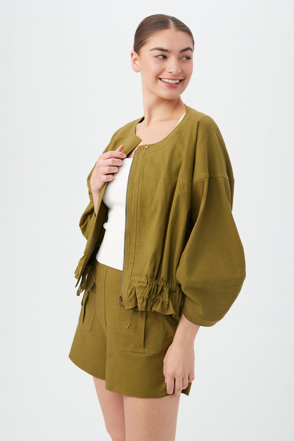 A woman with dark hair in a bun wears the olive green Prita Jacket with wide sleeves and matching shorts over a white sleeveless top. She smiles, looking left, standing against a plain light background.
