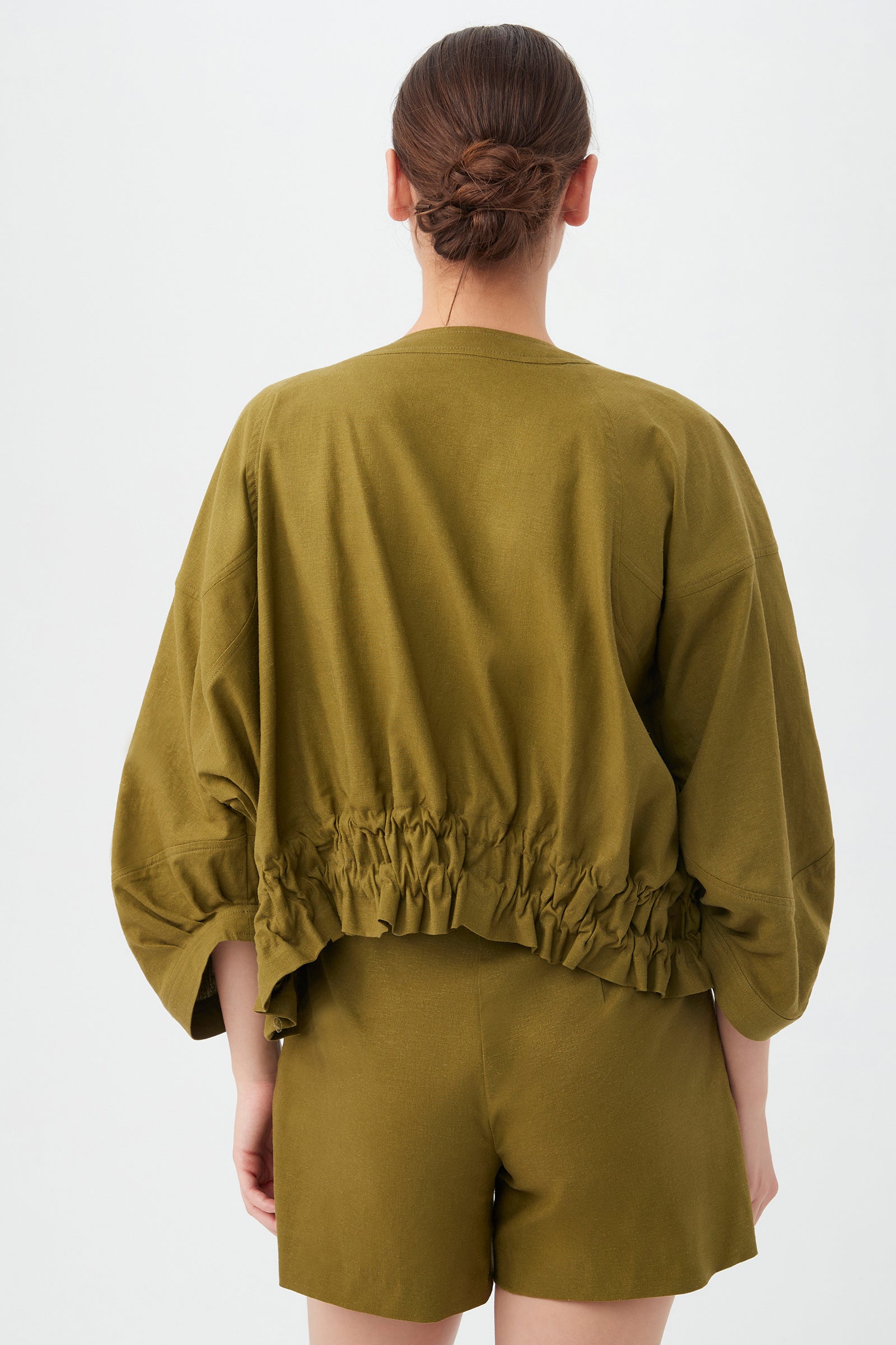 A woman with brown hair in a low bun is seen from the back, wearing the olive green Prita Jacket with a gathered waist and matching shorts, set against a plain white background.
