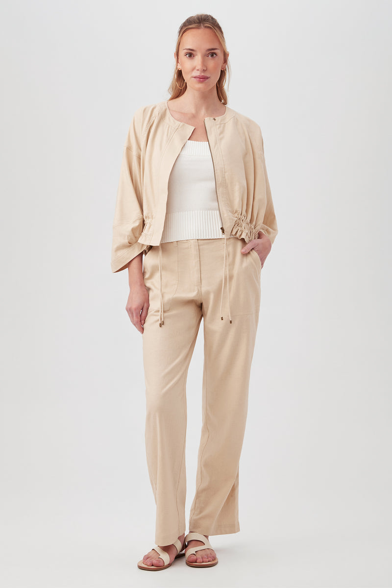 A woman stands facing forward, wearing the beige Prita Jacket with drawstrings, matching pants, a white ribbed top, and brown sandals. She has long light brown hair and a relaxed, confident expression.
