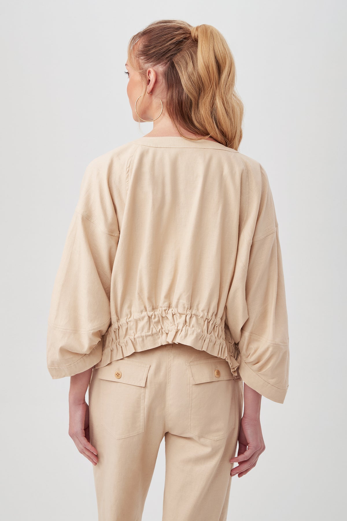 A woman with long, wavy blonde hair in a ponytail is shown from the back, wearing the Prita Jacket—a loose beige top with an elastic gathered waist and wide sleeves—paired with matching high-waisted pants.