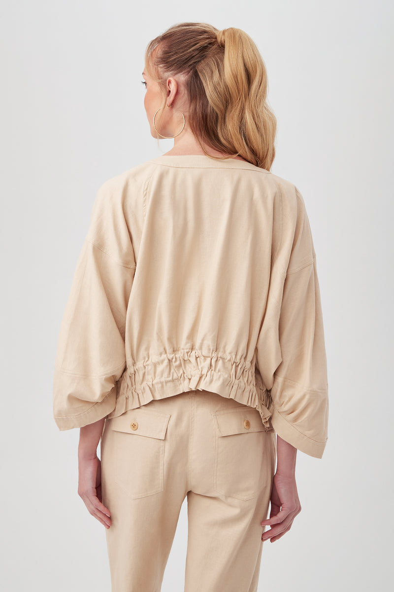 A woman with long, wavy blonde hair in a ponytail is shown from the back, wearing the Prita Jacket—a loose beige top with an elastic gathered waist and wide sleeves—paired with matching high-waisted pants.