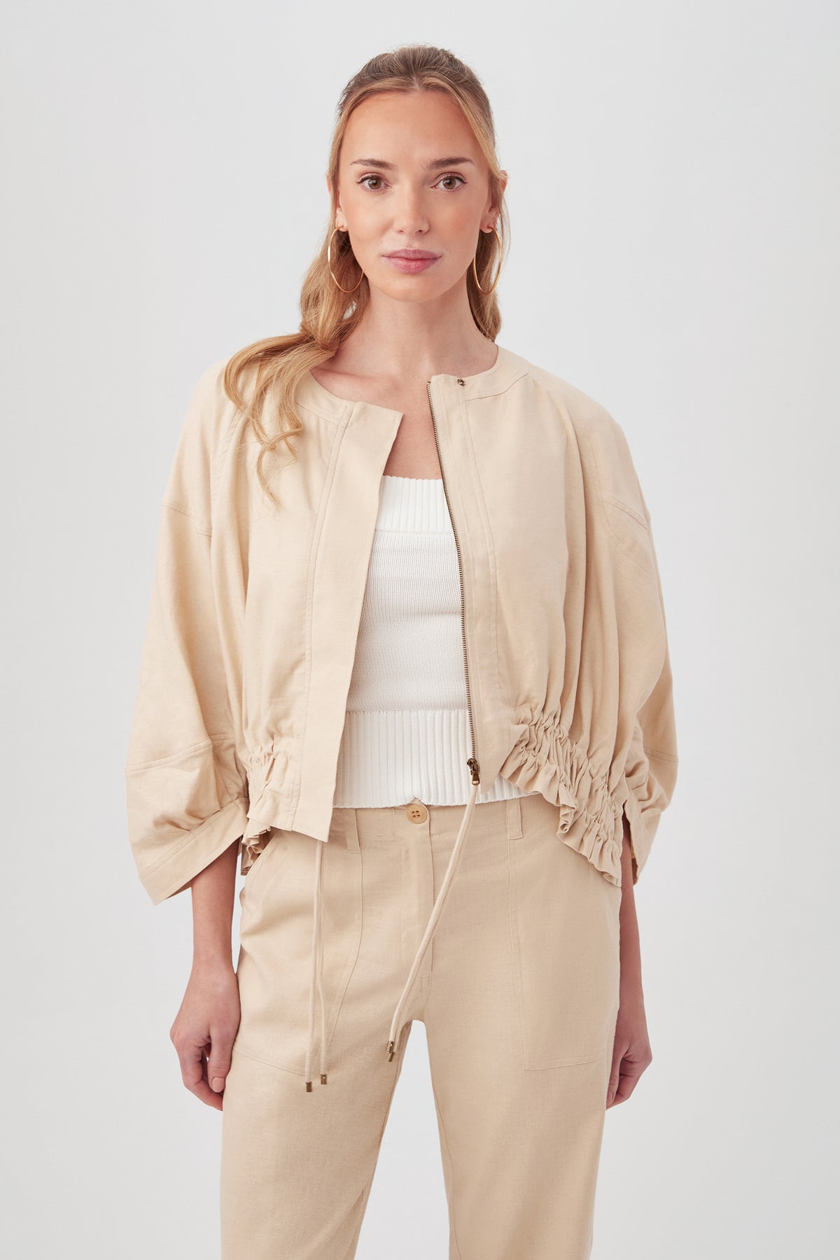A woman in the Prita Jacket in Biscotti, a light beige, loose-fitting style with drawstrings at the waist, layered over a white ribbed top and matching beige pants, stands against a plain light background.