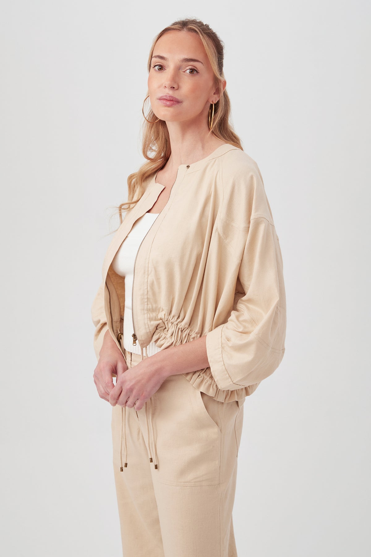 A woman with long, wavy blonde hair wears the beige Prita Jacket with matching pants over a white top, standing against a plain light background and looking slightly to the side with a calm expression.