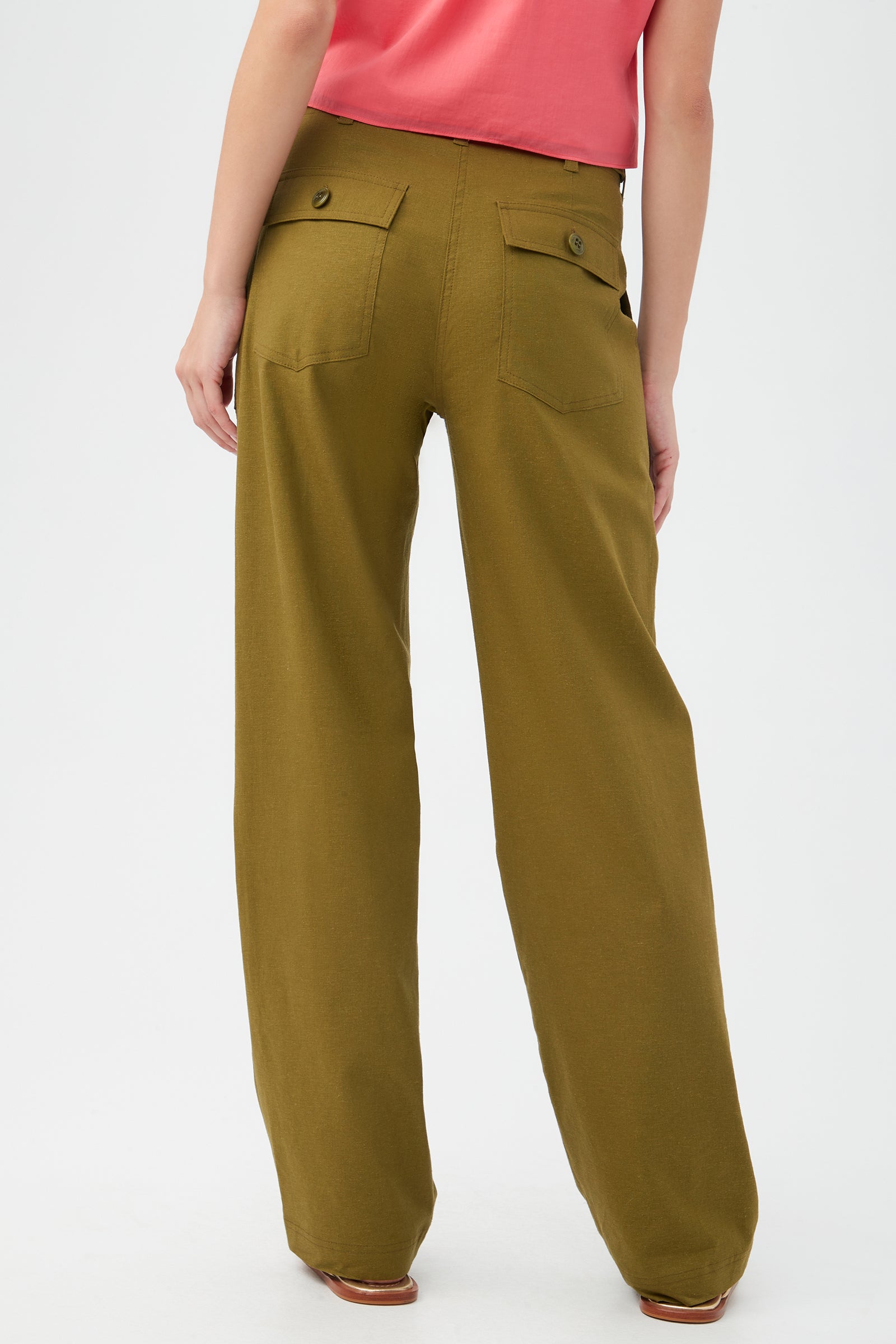 A person faces away, wearing the Antonia Pant in olive green—these wide-leg, loose-fitting pants have buttoned back pockets and are paired with a pink shirt and brown shoes. The background is plain and light-colored.
