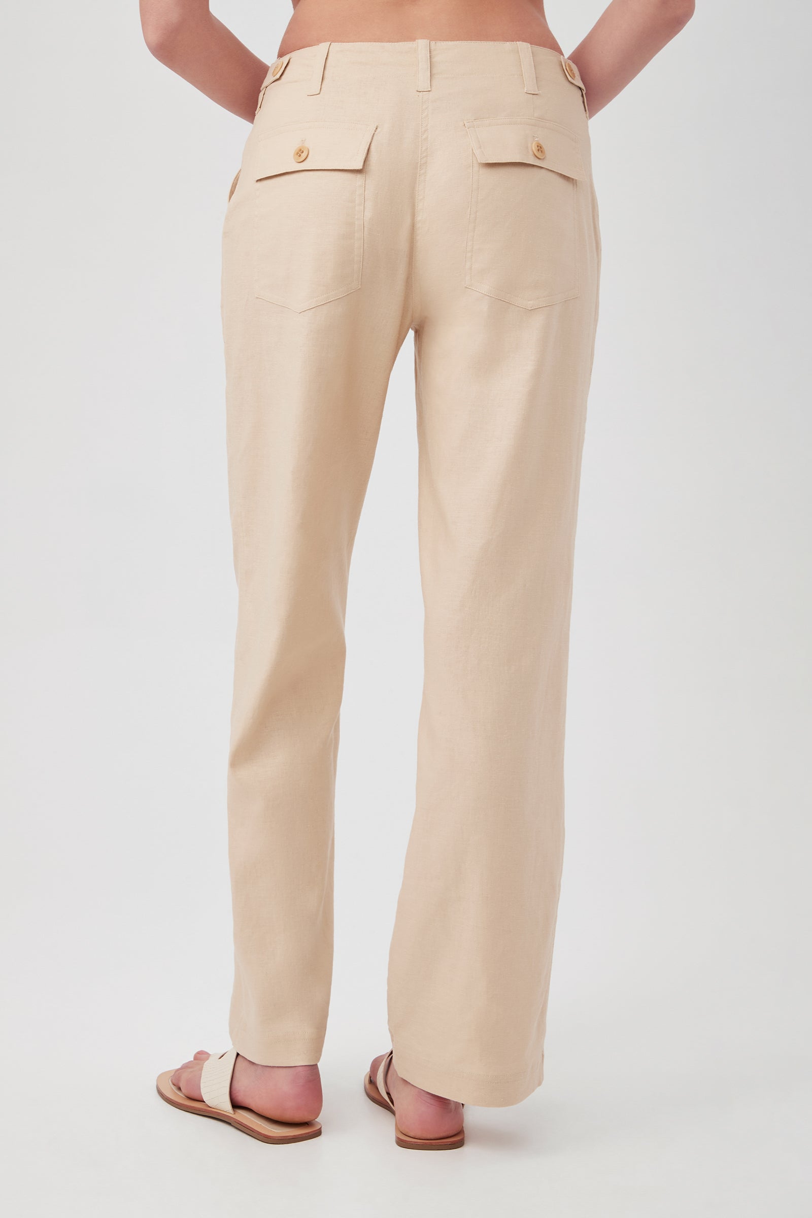 A person faces away, wearing the Antonia Pant in light beige with straight legs, two back pockets, tan buttons, and beige sandals. The background is plain white.