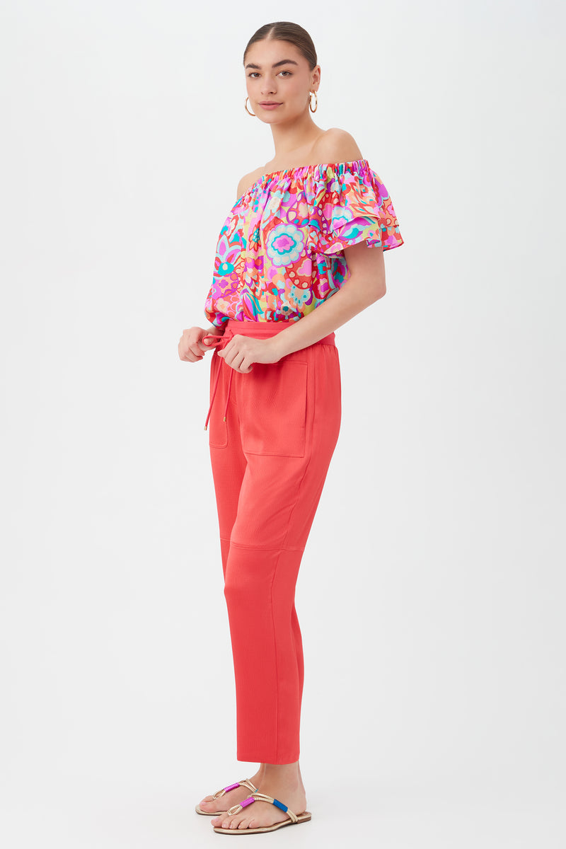 A woman stands sideways against a plain white background, wearing the Bernelle 2 Pant in coral with a colorful off-the-shoulder floral top, hoop earrings, and multicolored sandals.