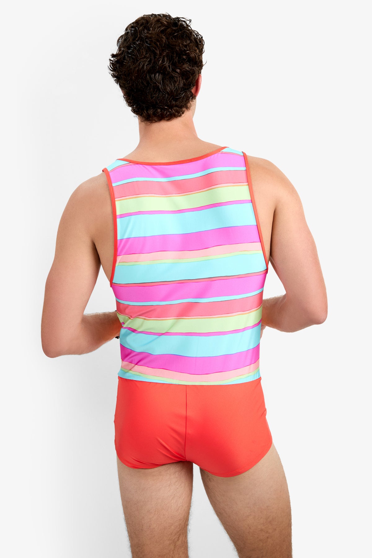 Seen from behind, a person with short curly hair wears the Sunset Beach Wetsuit - Sol Stripe, featuring vibrant horizontal stripes in pink, green, blue, and orange. The background is plain white.