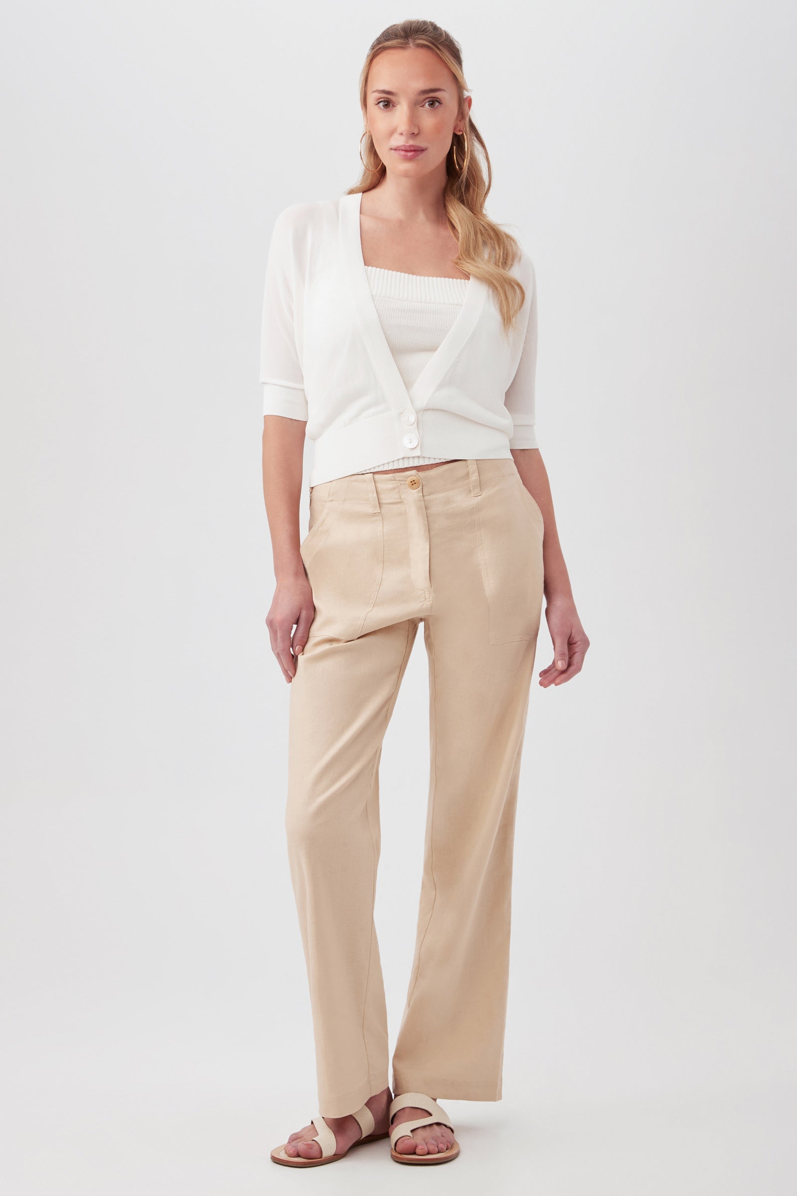 A woman poses confidently against a plain background, wearing beige high-waisted Antonia Pants, a white top with a matching cardigan, and beige sandals. Her loose waves hairstyle completes the relaxed look.