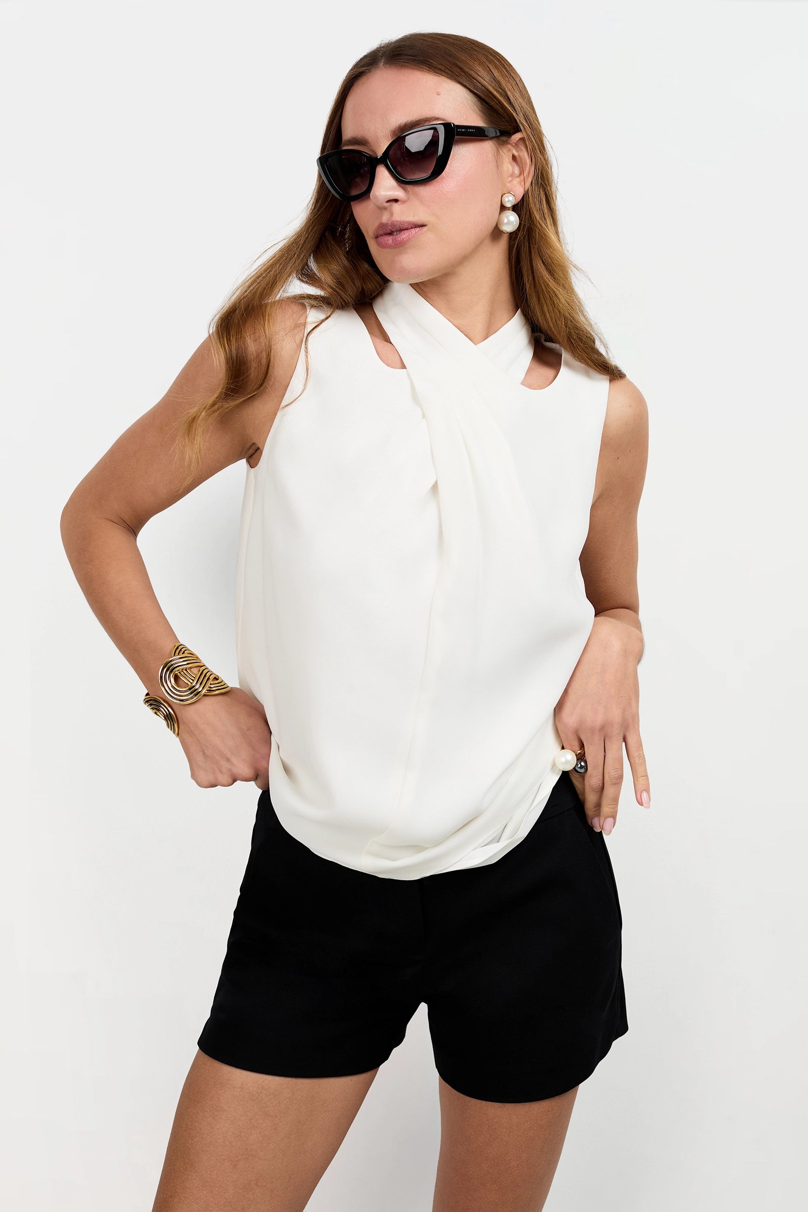 A woman models the Magni Cross-Neck Top—sleeveless and white—paired with black shorts, oversized black sunglasses, and gold jewelry against a plain white background.