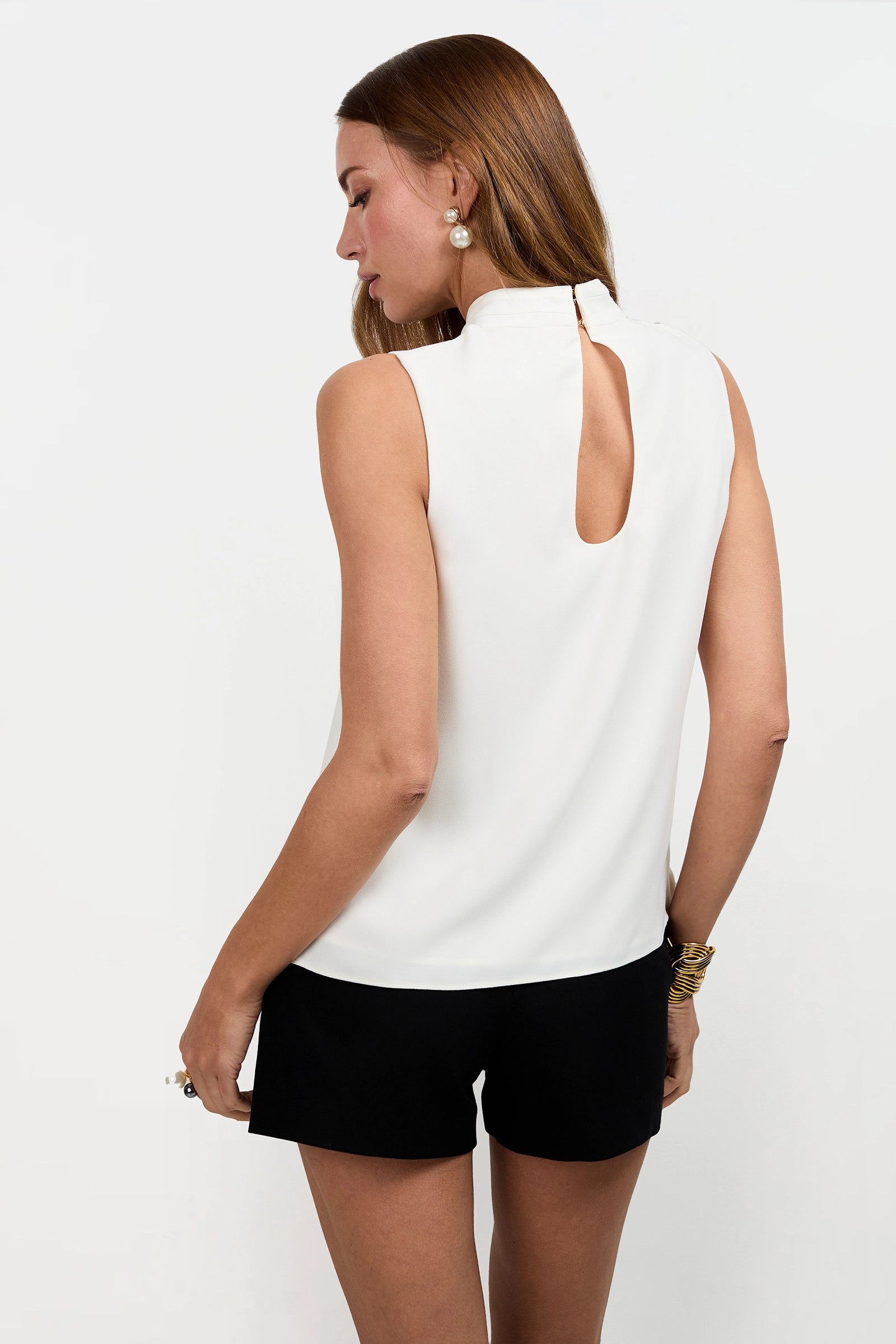A woman with long brown hair wears the Magni Cross-Neck Top—sleeveless and white with a keyhole back—paired with black shorts. She stands against a plain white background, slightly turned away to show the top's back details.