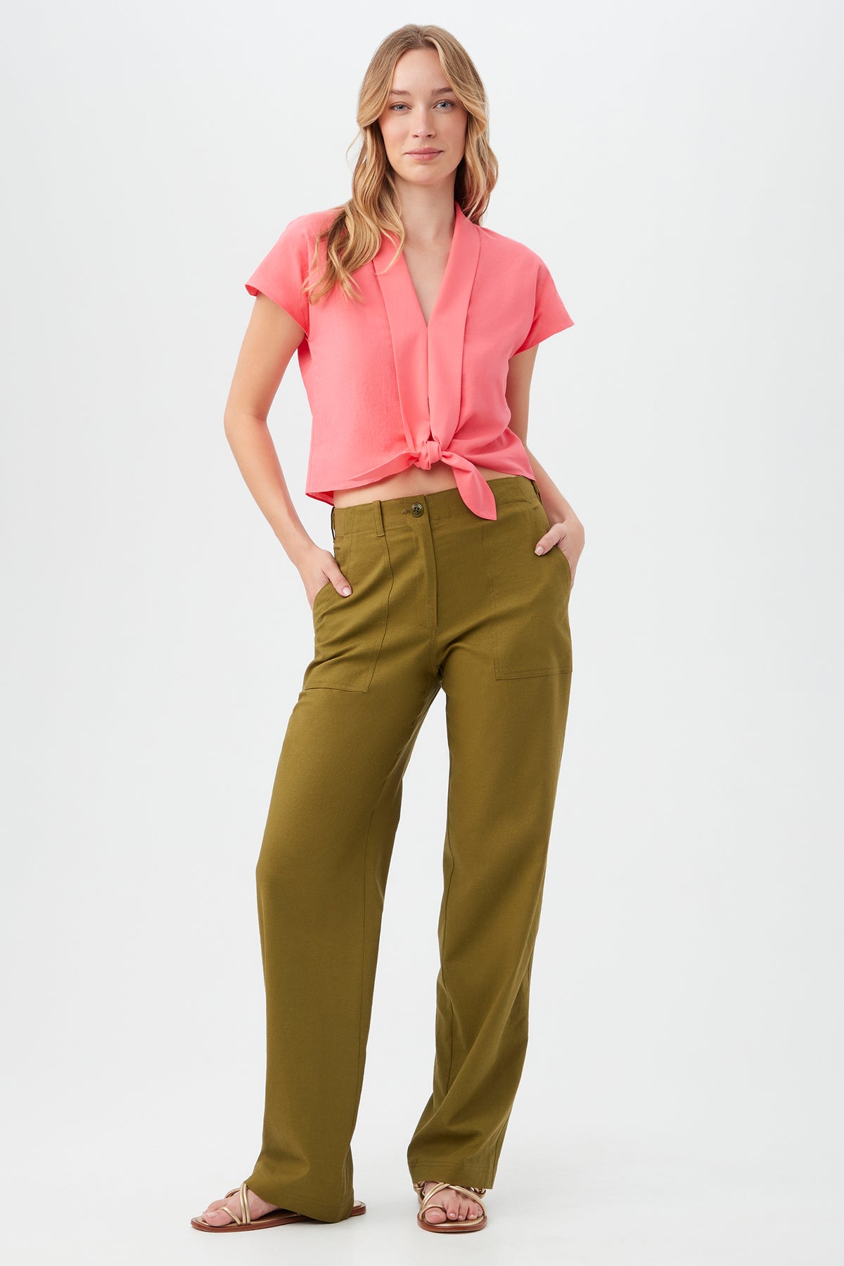 A woman with long, wavy hair poses confidently against a plain background, wearing high-waisted olive green Antonia Pants with a short-sleeve pink tie-front blouse and flat sandals.