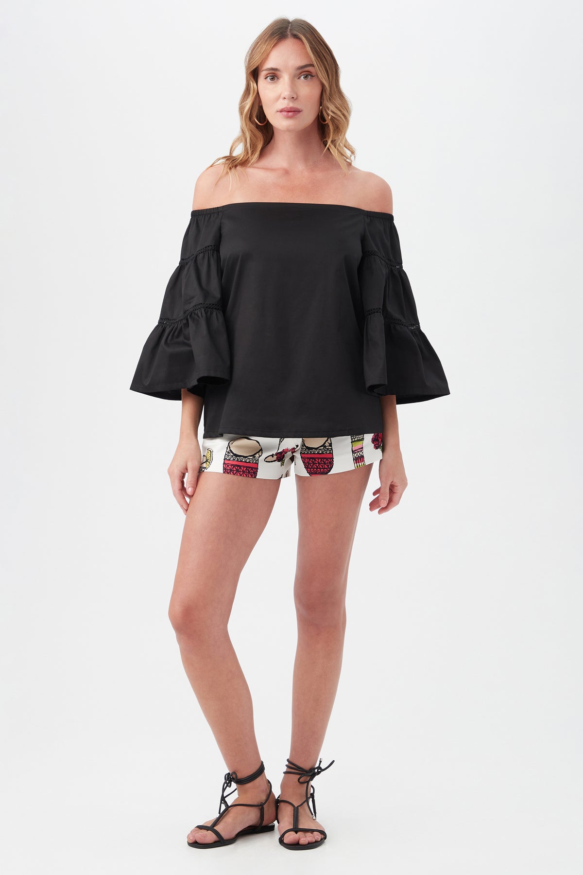 A woman stands against a white background wearing the Garnish Top, an off-the-shoulder black blouse with ruffled sleeves, paired with patterned shorts and black lace-up sandals.