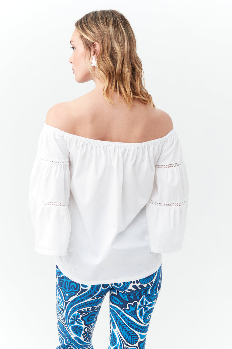 A woman with wavy blonde hair faces away, wearing the Garnish Top—an off-the-shoulder white blouse with long sleeves—and blue pants with a bold paisley pattern on a white background.