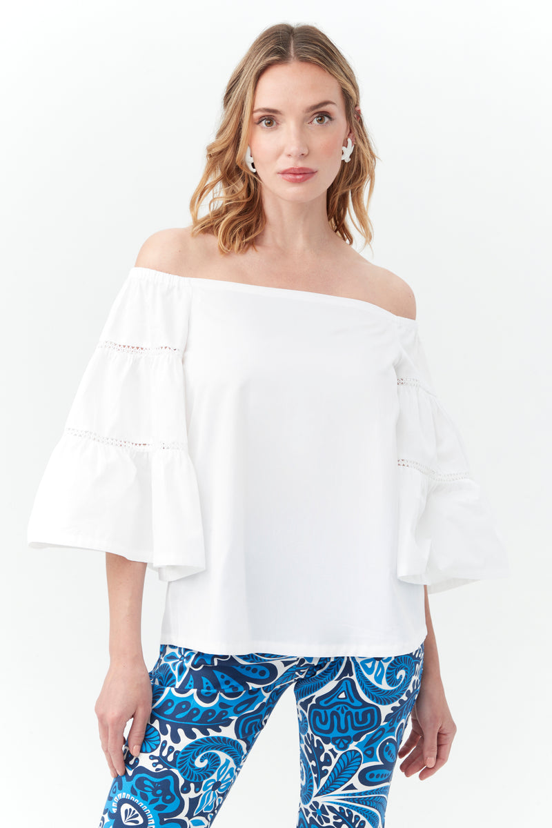 A woman models the white Garnish Top, an off-the-shoulder blouse with bell sleeves, paired with blue patterned pants, standing against a plain white background.