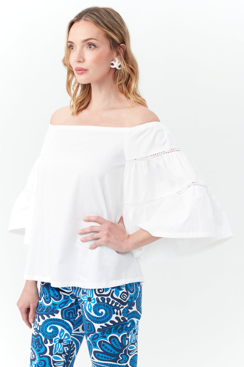 A woman wears the Garnish Top, a white off-the-shoulder blouse with ruffled sleeves, paired with blue patterned pants, white flower earrings, and stands against a plain white background. She has wavy light brown hair.