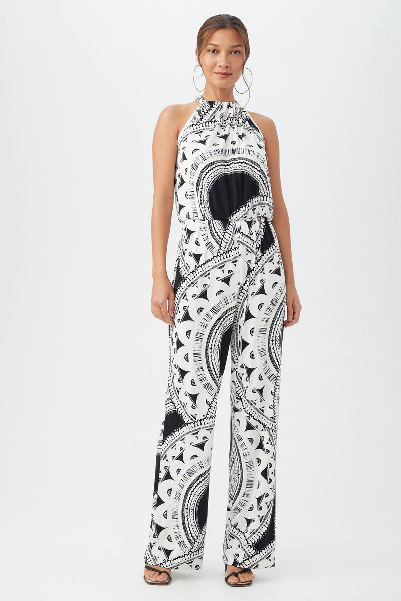 A woman poses against a white background wearing the Rancho Jumpsuit in multi, featuring a sleeveless design, high neck, wide-leg pants, and styled with hoop earrings and open-toe sandals.