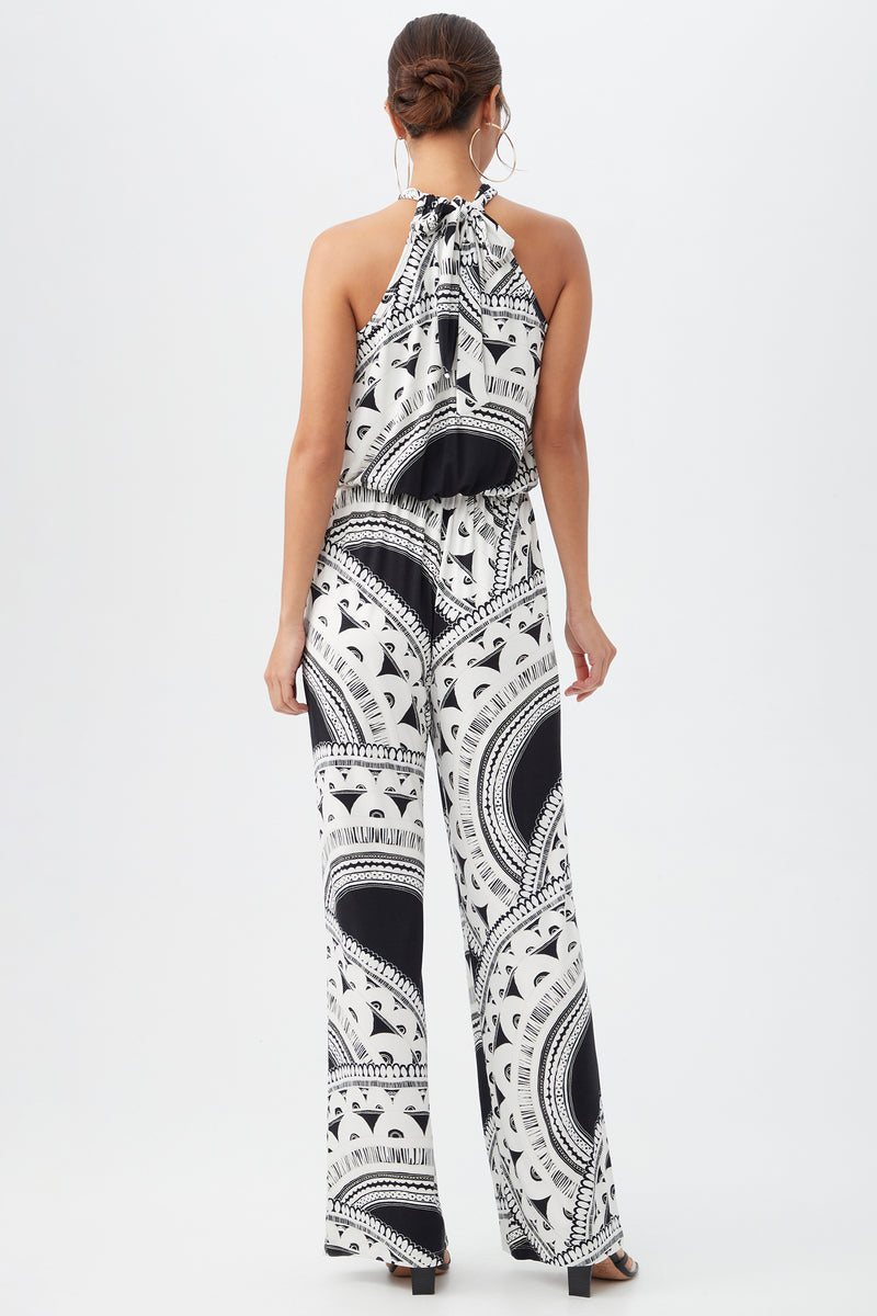 A woman stands against a white background, wearing the Rancho Jumpsuit—a sleeveless black and white patterned halter style with wide legs and an open back. She pairs it with hoop earrings, a low bun hairstyle, and black heels.