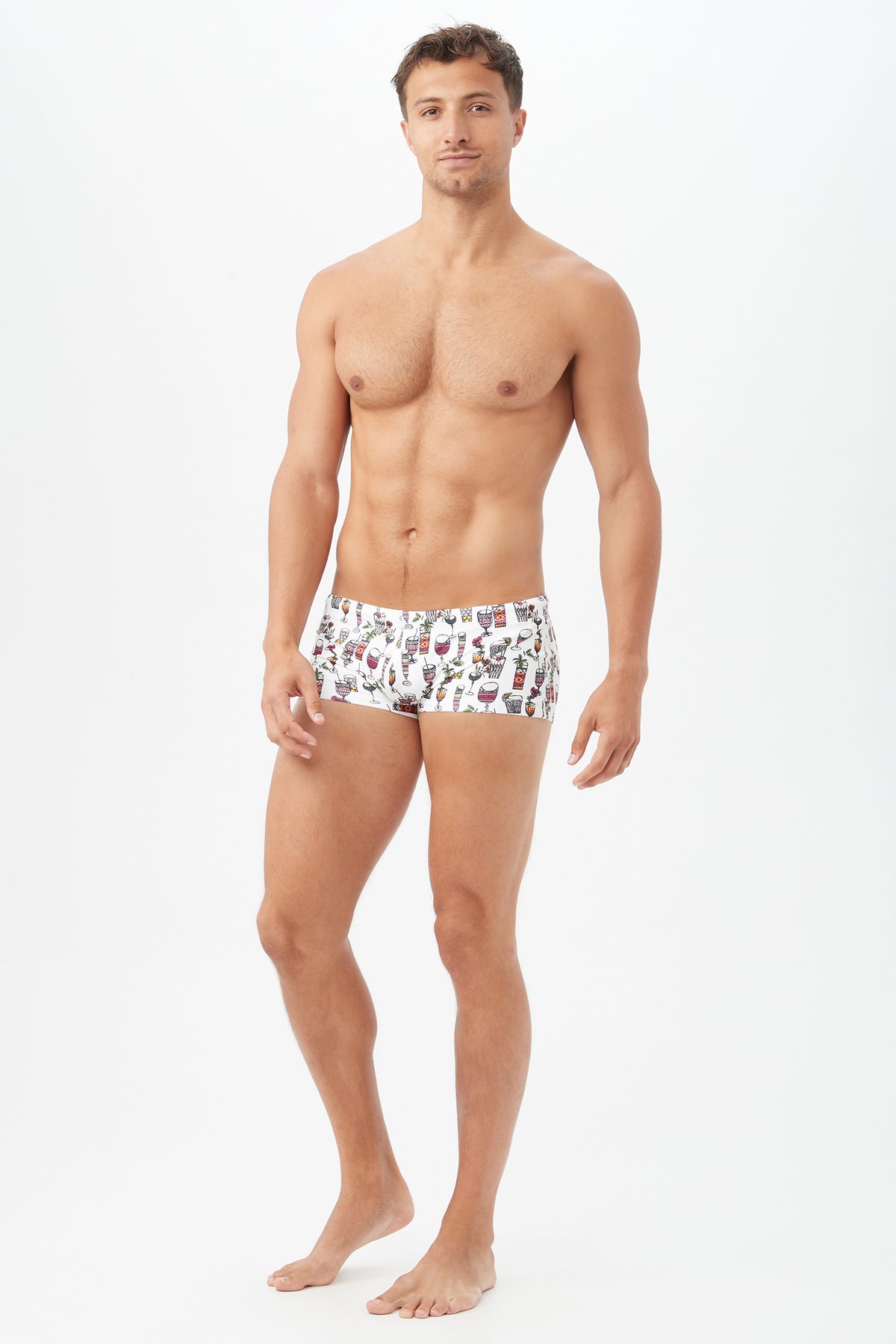 A man stands barefoot against a white background, facing forward with a neutral expression, wearing the Sorrento Swim Trunk – Happy Hour Swim Knit, featuring a vibrant and playful pattern.