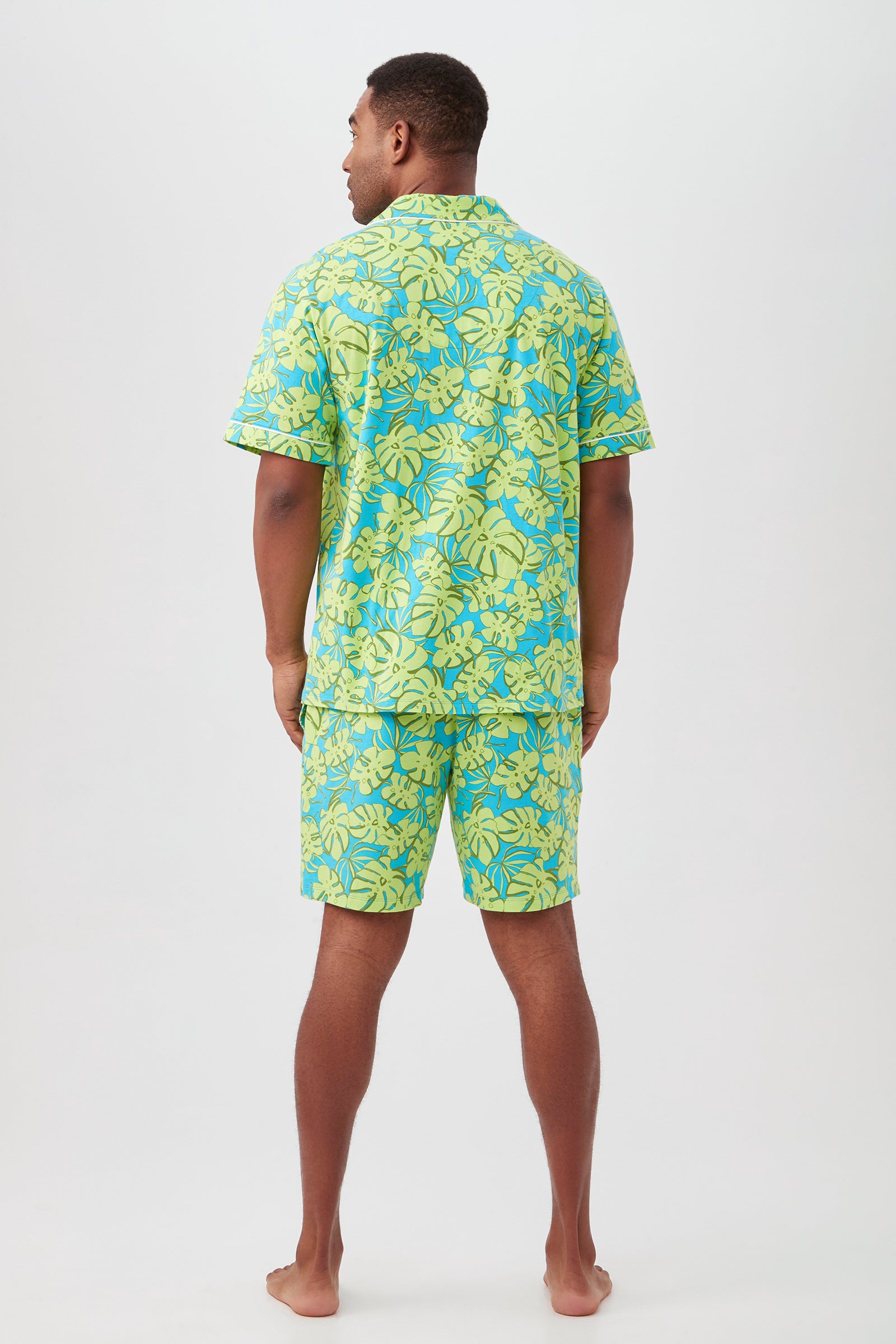 A man stands barefoot, facing away from the camera, modeling the Palm Leaves Mens Short Sleeve Boxer Short Jersey PJ Set with a blue and green tropical leaf pattern, against a plain white background.