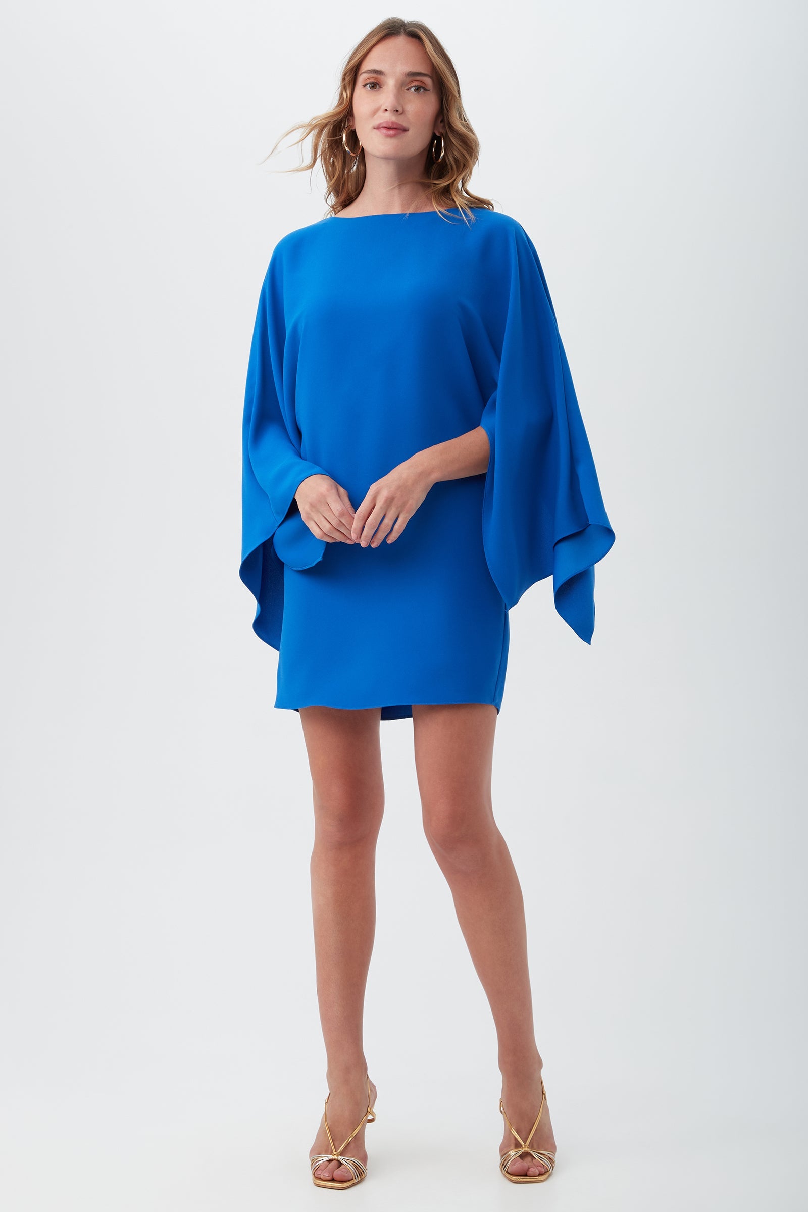 A woman with light brown, wavy hair wears the Daiquiri Dress, a vibrant royal blue mini dress featuring wide batwing sleeves. She pairs it with gold open-toe heels and stands against a plain background.