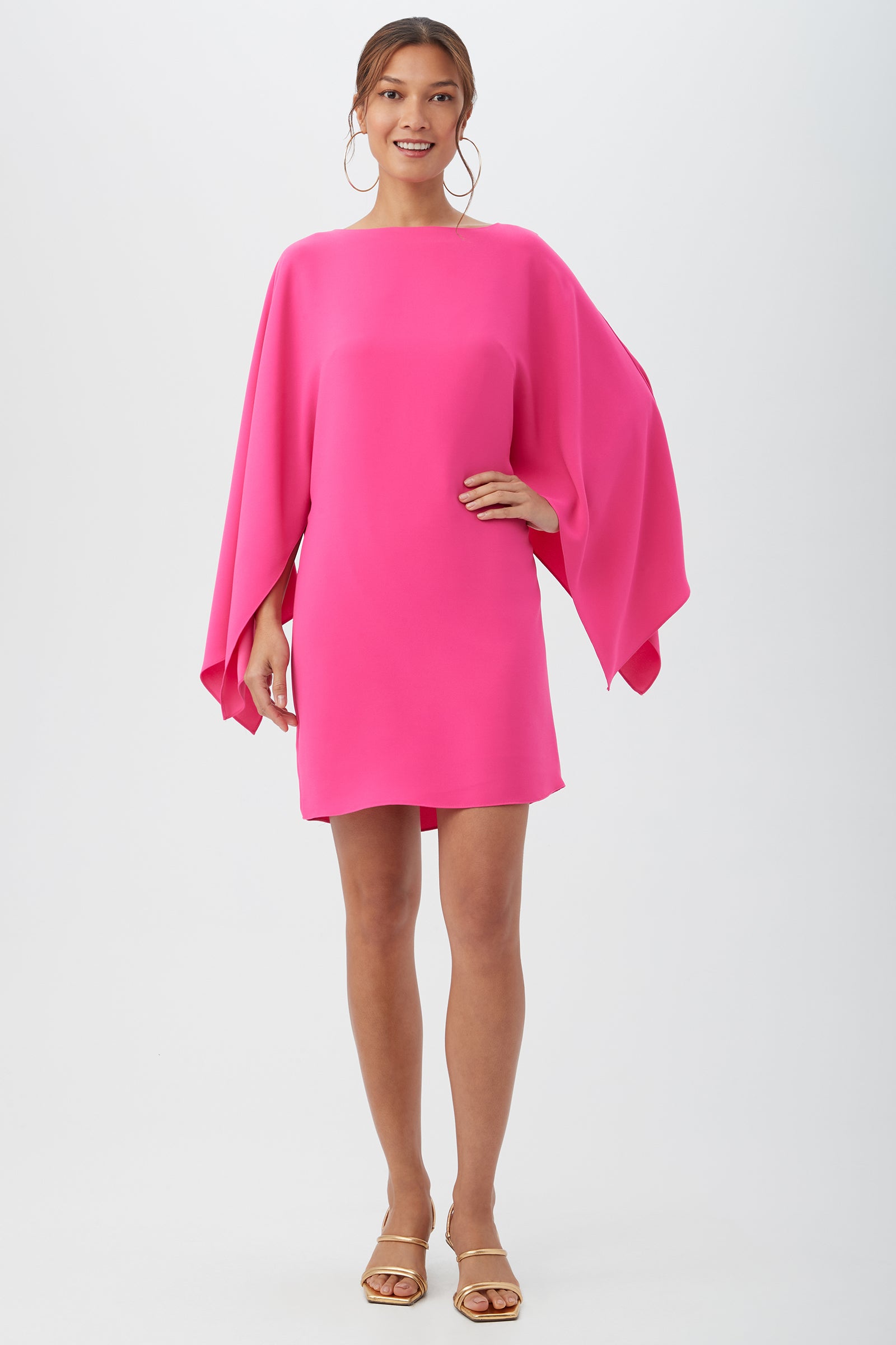 A woman smiles in the loose-fitting Daiquiri Dress in Lillikoi, featuring cape sleeves. She accessorizes with large hoop earrings and strappy nude sandals against a plain white background.