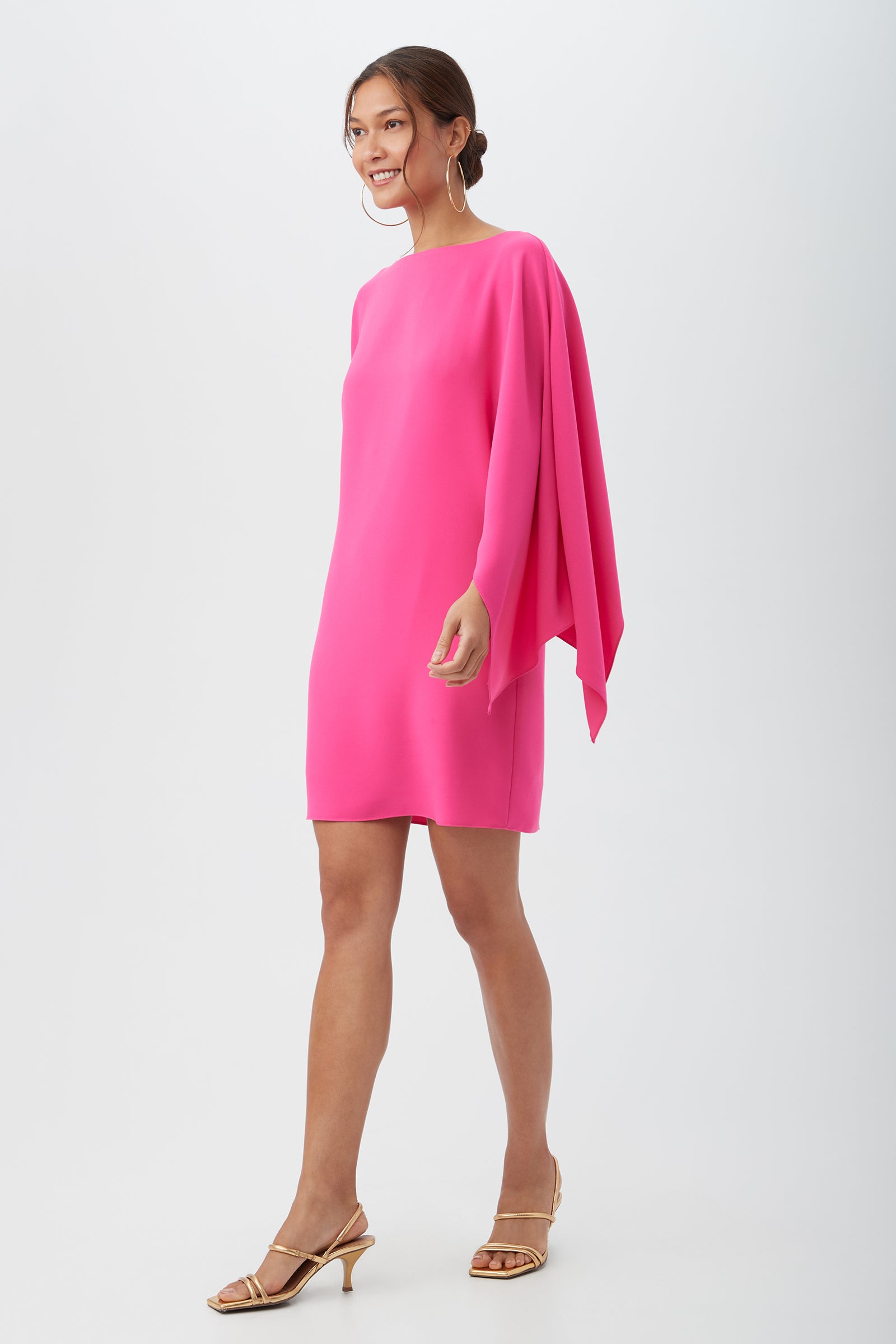 A woman smiles in the Daiquiri Dress, a bright pink, knee-length style featuring a cape-like sleeve on one side. She accessorizes with gold hoop earrings and strappy gold heels, her hair in a low bun against a white background.