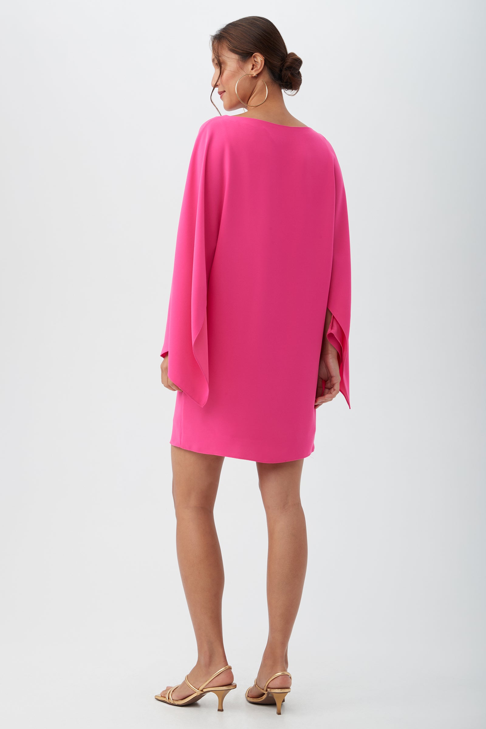 A woman with her hair in a low bun and large hoop earrings stands facing away, wearing the Daiquiri Dress—a bright pink, loose-fit style with wide sleeves—and beige heeled sandals against a plain background.