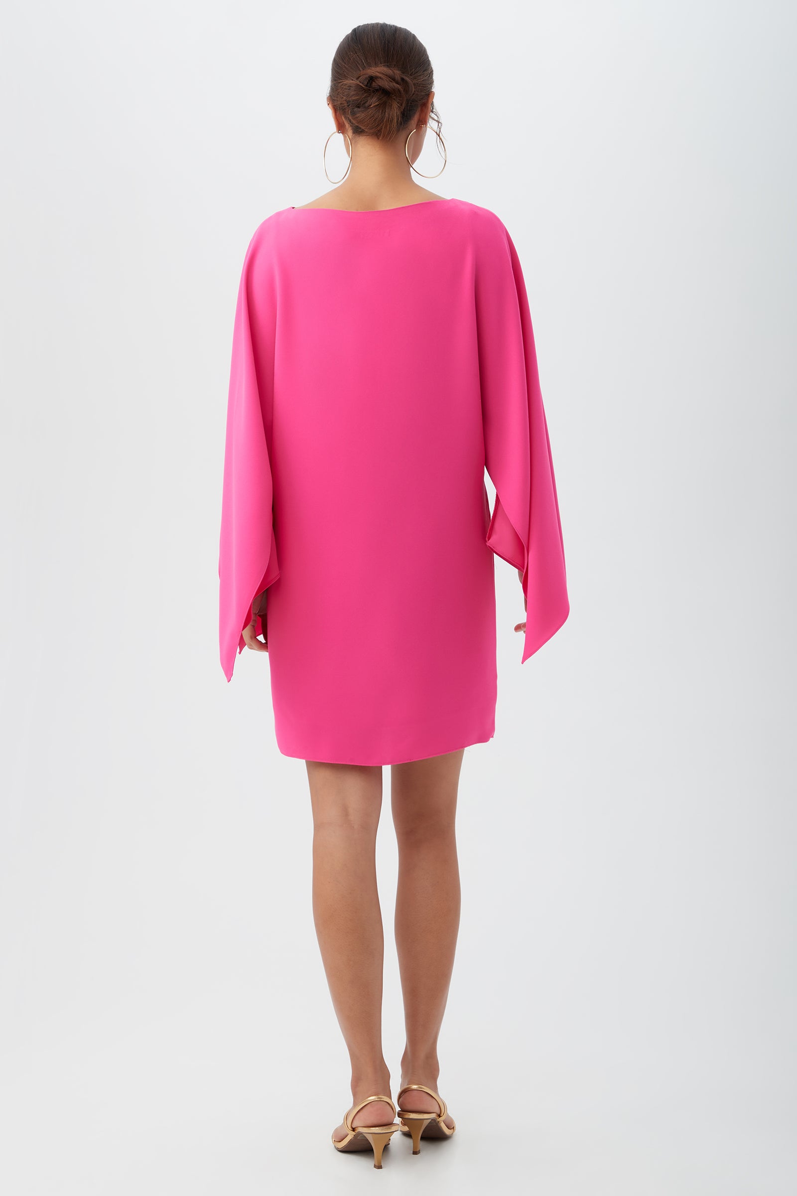 A woman faces away, wearing the Daiquiri Dress in bright pink with flowing sleeves and beige heeled sandals, set against a plain white background.