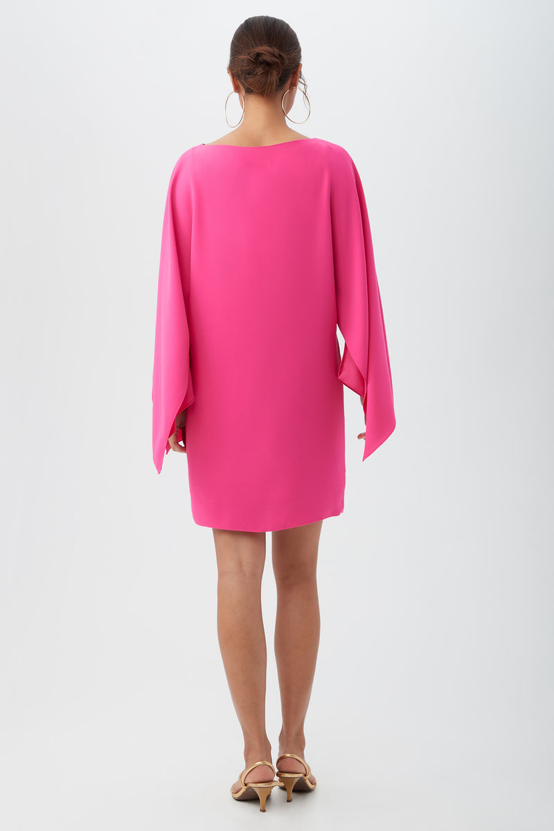 A woman faces away, wearing the Daiquiri Dress in bright pink with flowing sleeves and beige heeled sandals, set against a plain white background.