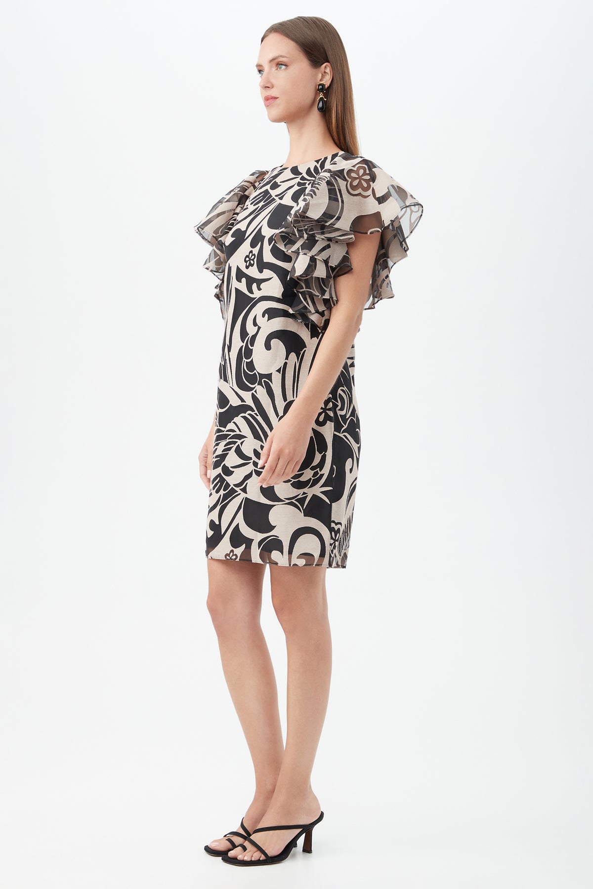 Woman in profile wears the Hokulani Dress, a knee-length black and beige floral print with layered ruffle sleeves, paired with black heeled sandals and statement earrings, set against a plain white background.