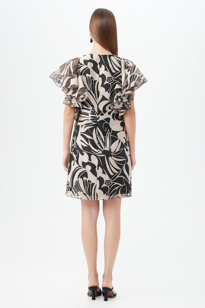 A woman faces away, wearing the Hokulani Dress—a short black-and-tan floral dress with flutter sleeves—and black heeled sandals. Her long, straight hair is loose, set against a plain white background.