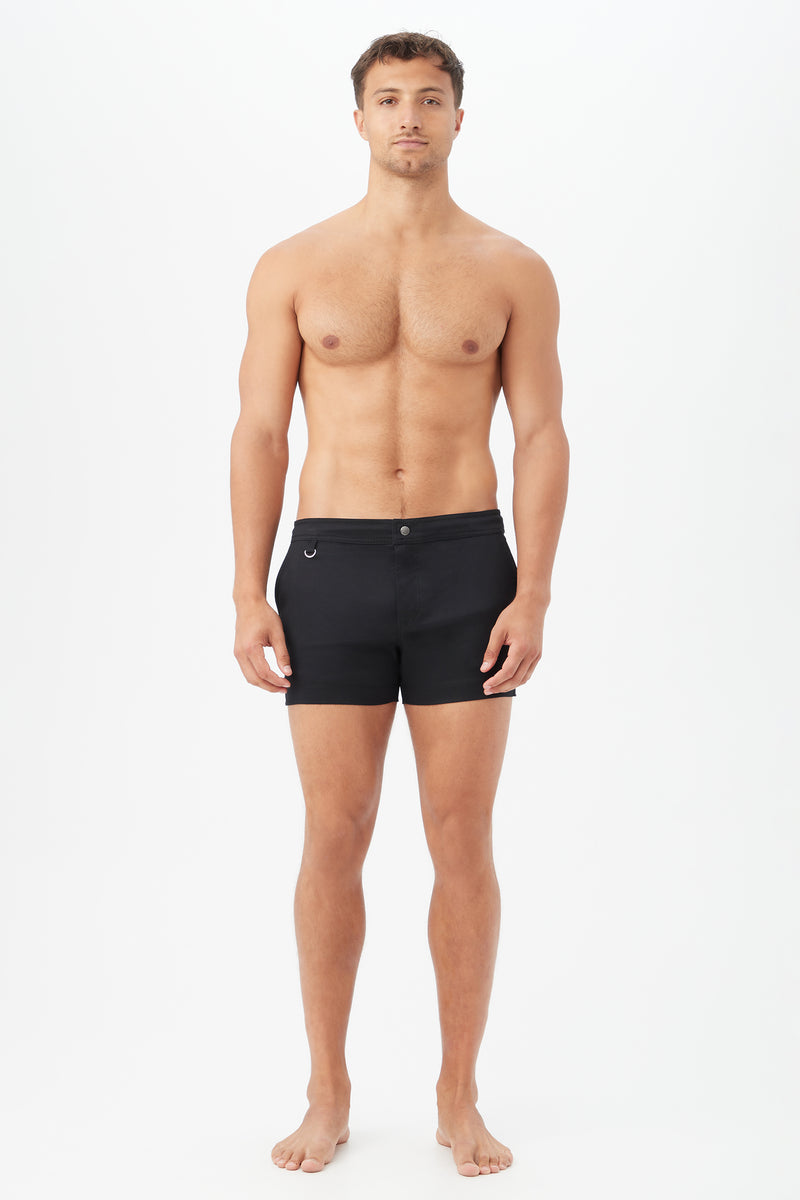 A man with short brown hair stands barefoot against a plain white background, facing forward in a neutral pose, wearing the Chico Swim Trunk - Metallic Swim Woven in black.