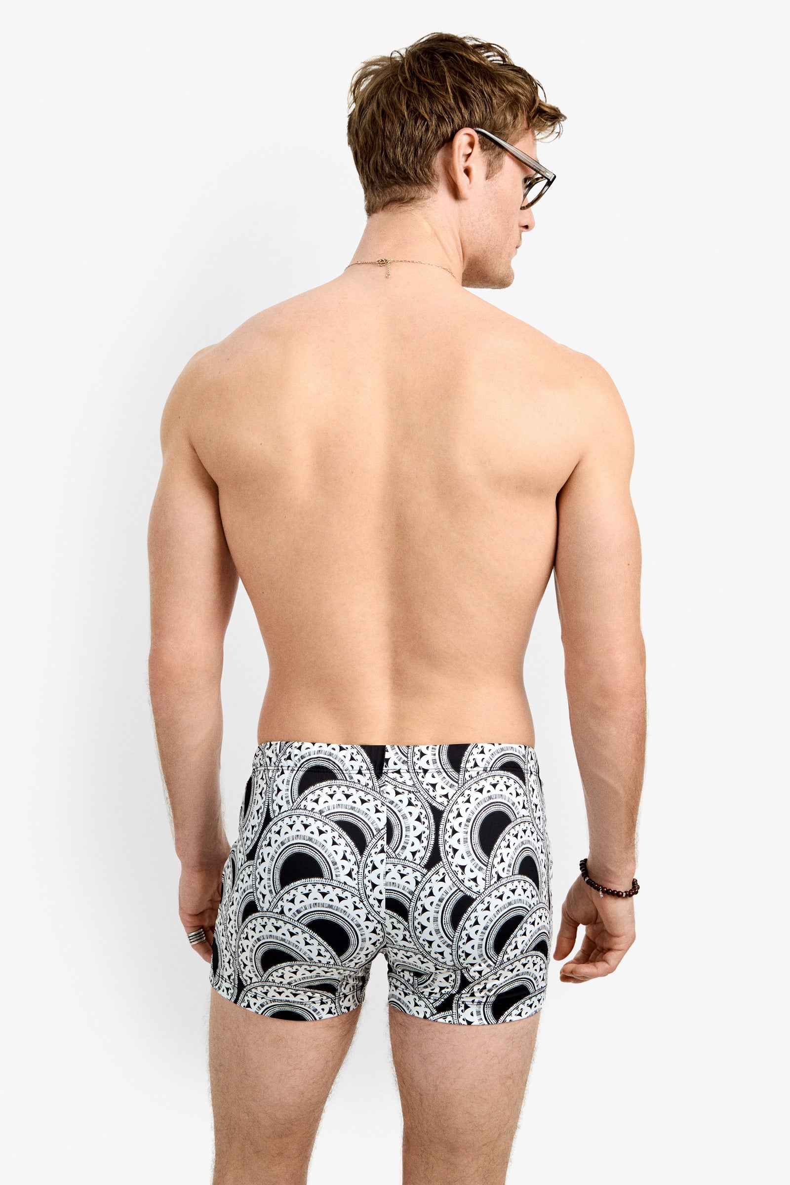 A man with short hair and glasses, seen from behind, stands against a plain white background wearing the Palm Desert Swim Trunk and a bracelet.