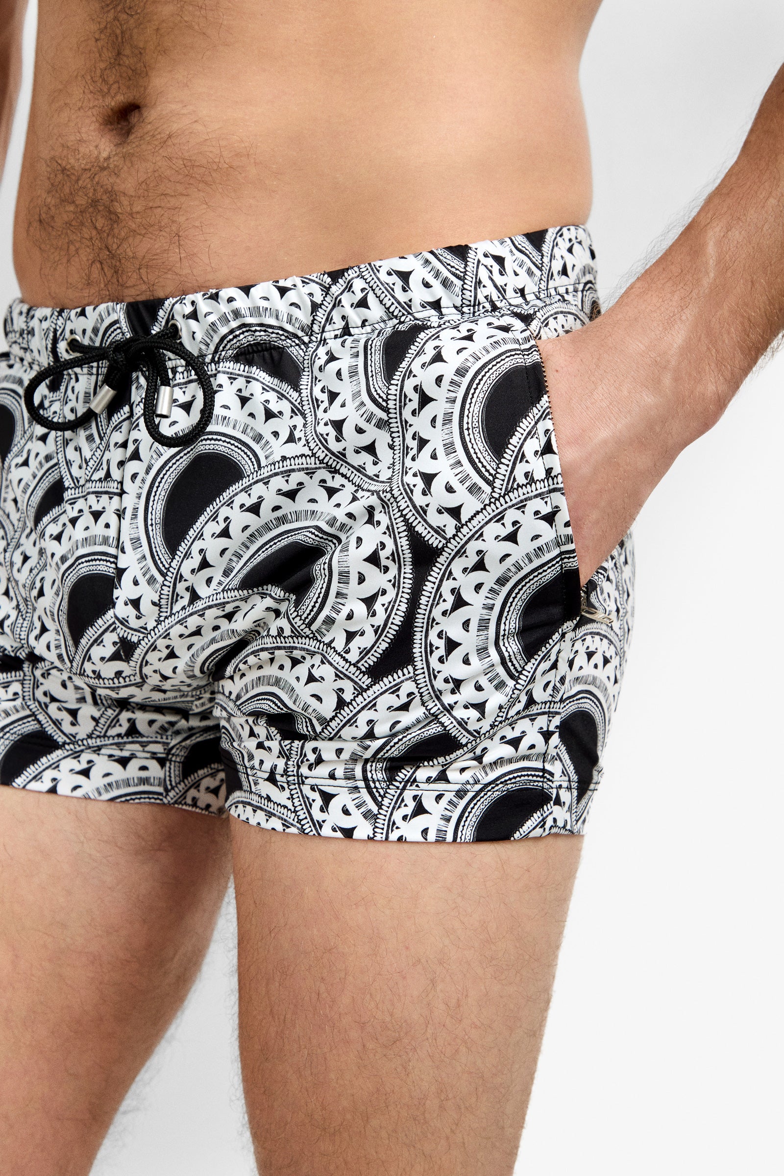A close-up shows a person wearing Palm Desert Swim Trunk, featuring black and white circular patterns and a drawstring, with one hand in the pocket. Only the person's torso and upper thighs are visible.