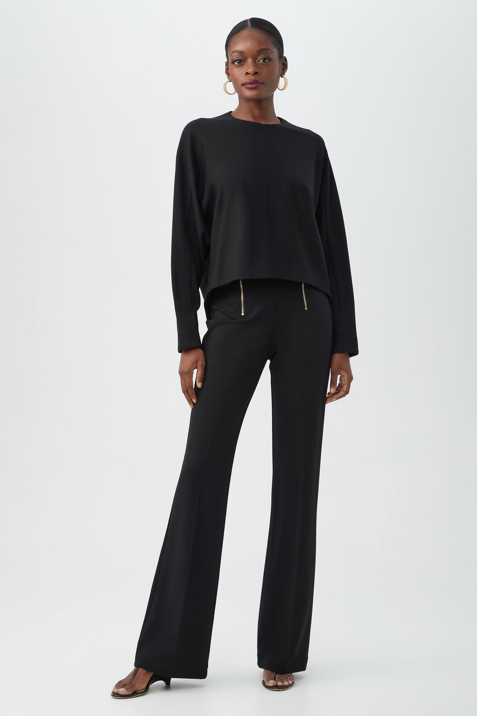 A woman stands against a plain background wearing the black Kiara Pant with zipper details, a loose black long-sleeve top, black heeled sandals, and gold hoop earrings.