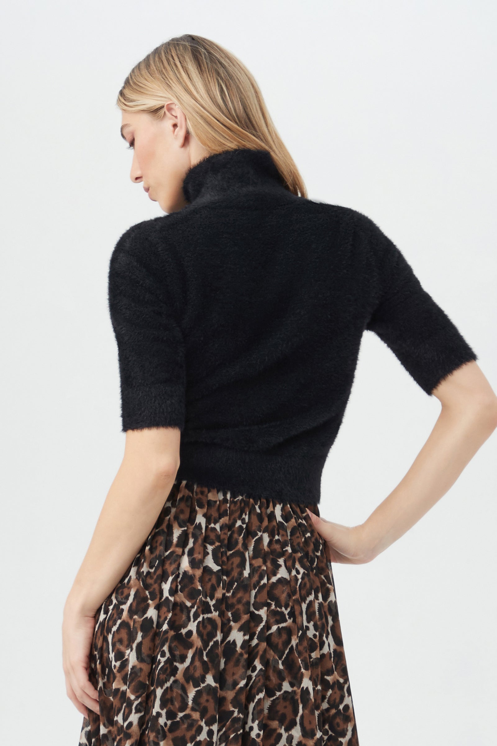 A woman with blonde hair, seen from behind, wears the Mali Fuzzy Sweater—a black short-sleeve turtleneck—paired with a high-waisted brown leopard print skirt against a plain white background.