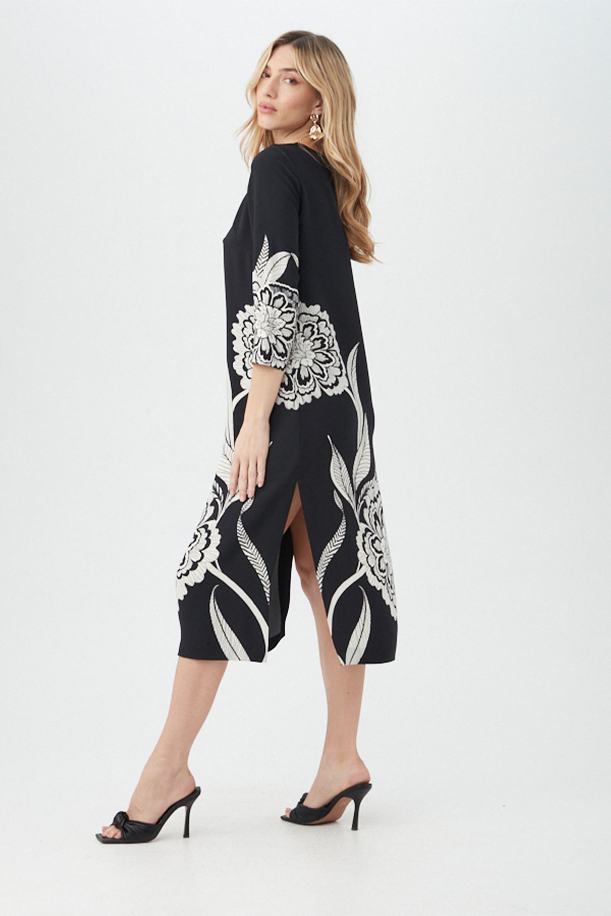 A woman models the Cointreau 2 Dress, a black knee-length dress with bold white floral patterns. She poses sideways on a plain white background, wearing black high-heeled sandals and gold earrings, with one hand on her waist.