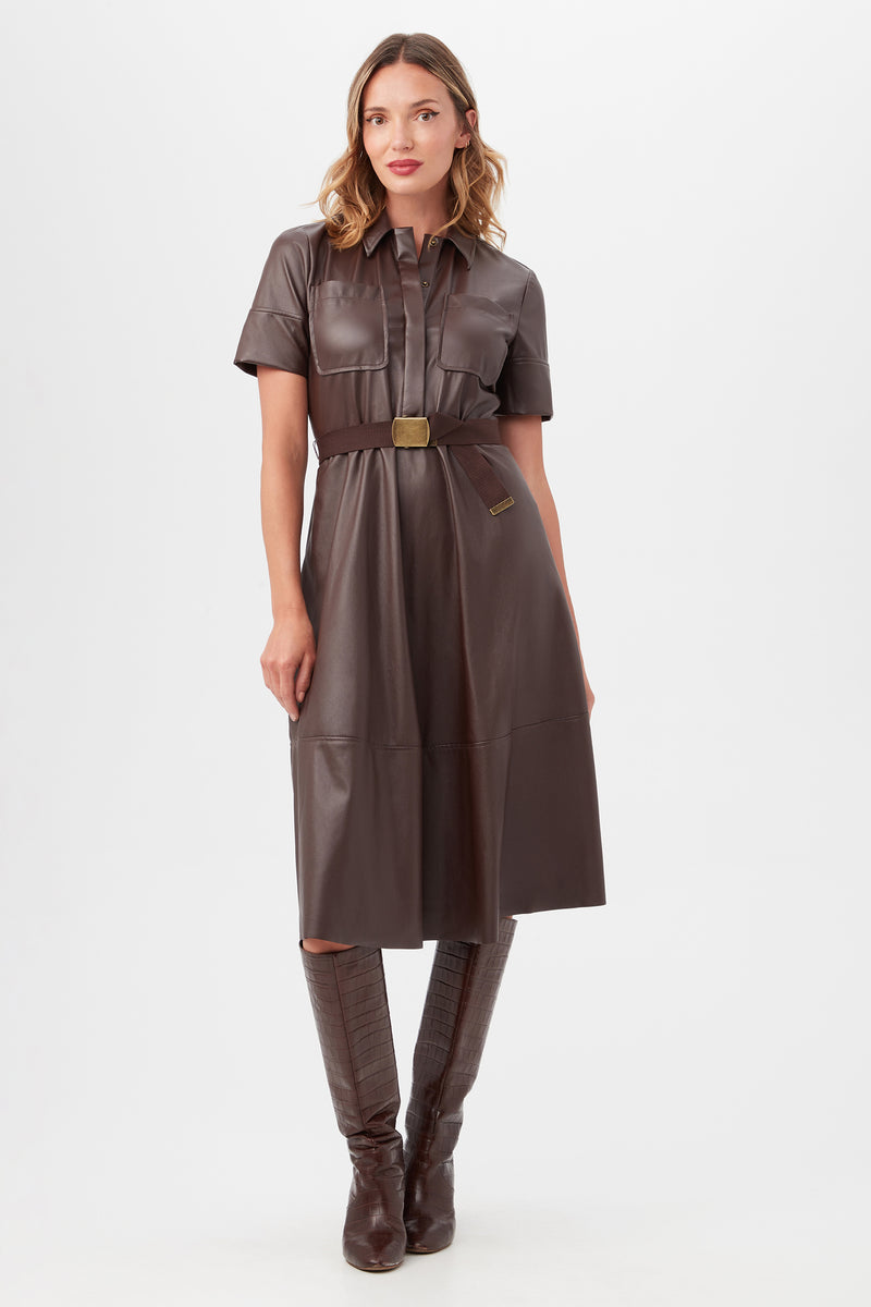 A woman with shoulder-length wavy hair models the Misola Dress in Mudcloth, a brown faux leather, short-sleeve, knee-length dress with a belted waist. She pairs it with tall brown boots against a plain white background.